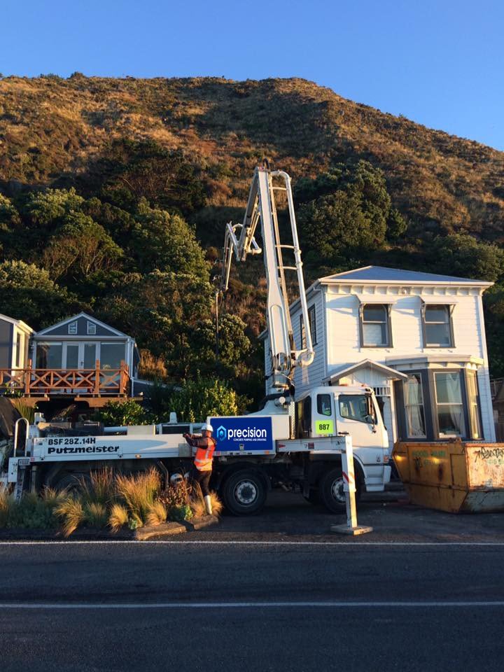 A Precision Concrete Pumping and Spraying  concrete pump with a boom lift parked on the side of a road, with a worker in a safety vest working nearby. Behind it are houses and a hillside covered in trees and shrubs.