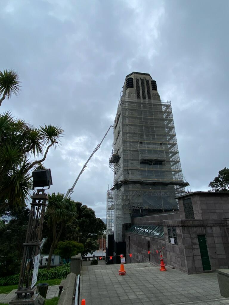 A tall building under construction covered with scaffolding, with a crane extending towards the top. The sky is cloudy, and there are trees and construction cones in the foreground.