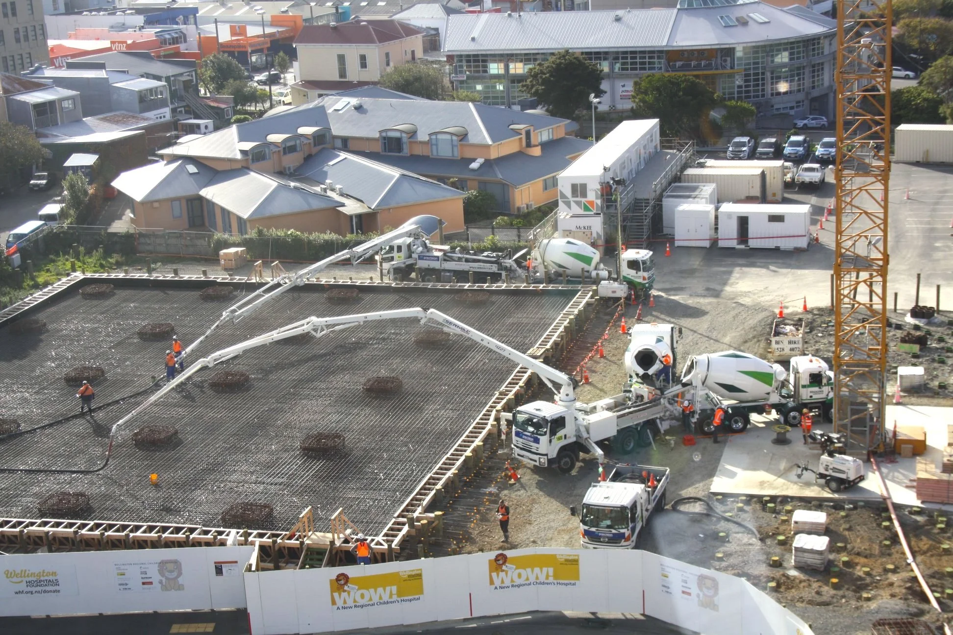 Construction workers project a concrete slab at a building site, with surrounding construction equipment and parked vehicles.