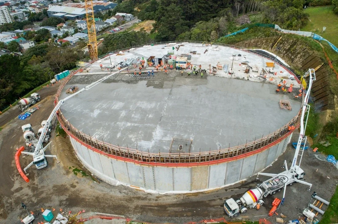 A construction site on top of a large circular concrete structure, with workers and construction equipment including cranes, cement mixers, and safety barriers, surrounded by residential areas and green hills.