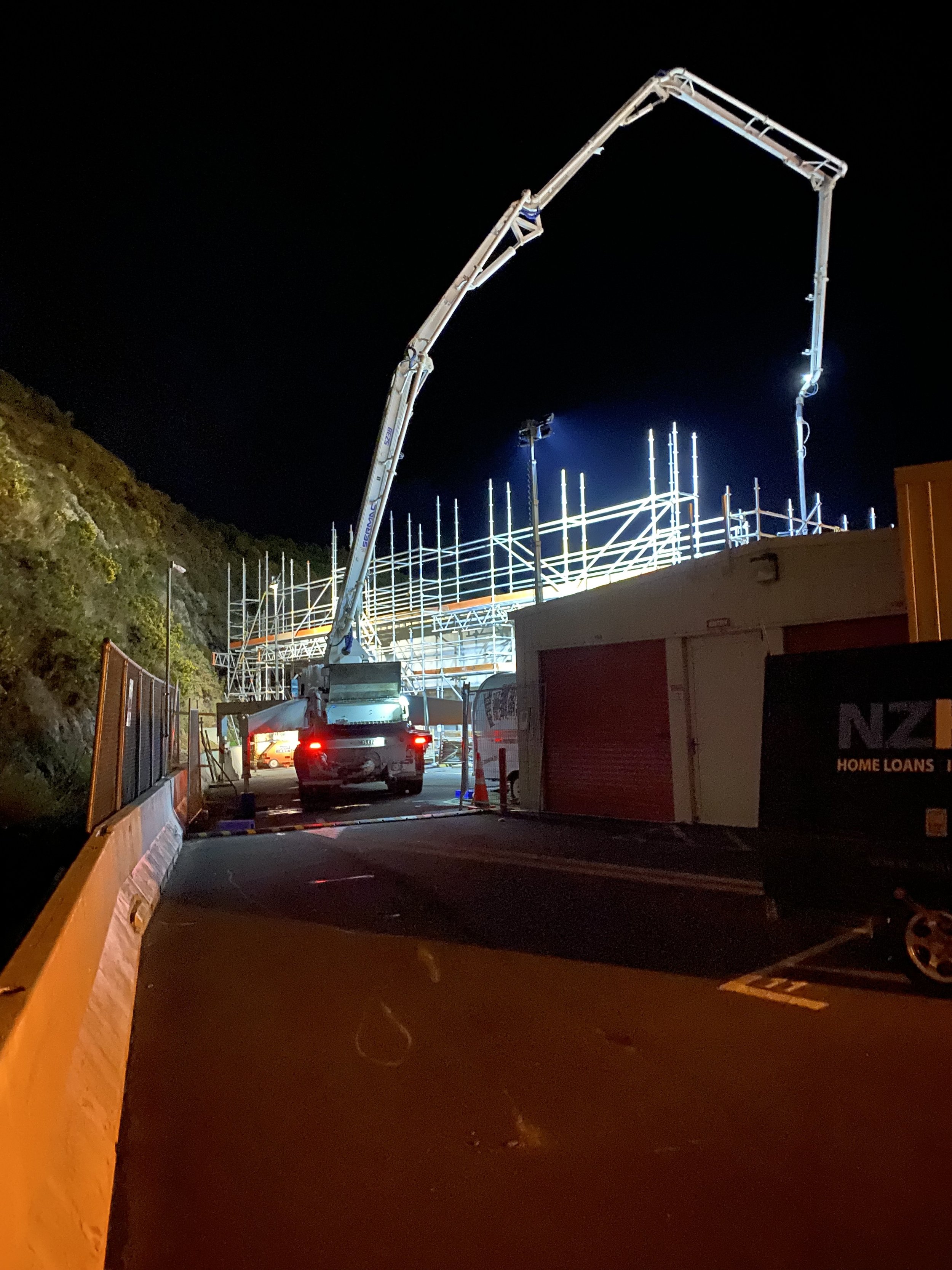 Nighttime construction site with scaffolding and a Precision Concrete Pumping and Spraying boom pump pumping concrete, illuminated by lights, on a hillside with a parked vehicle nearby.