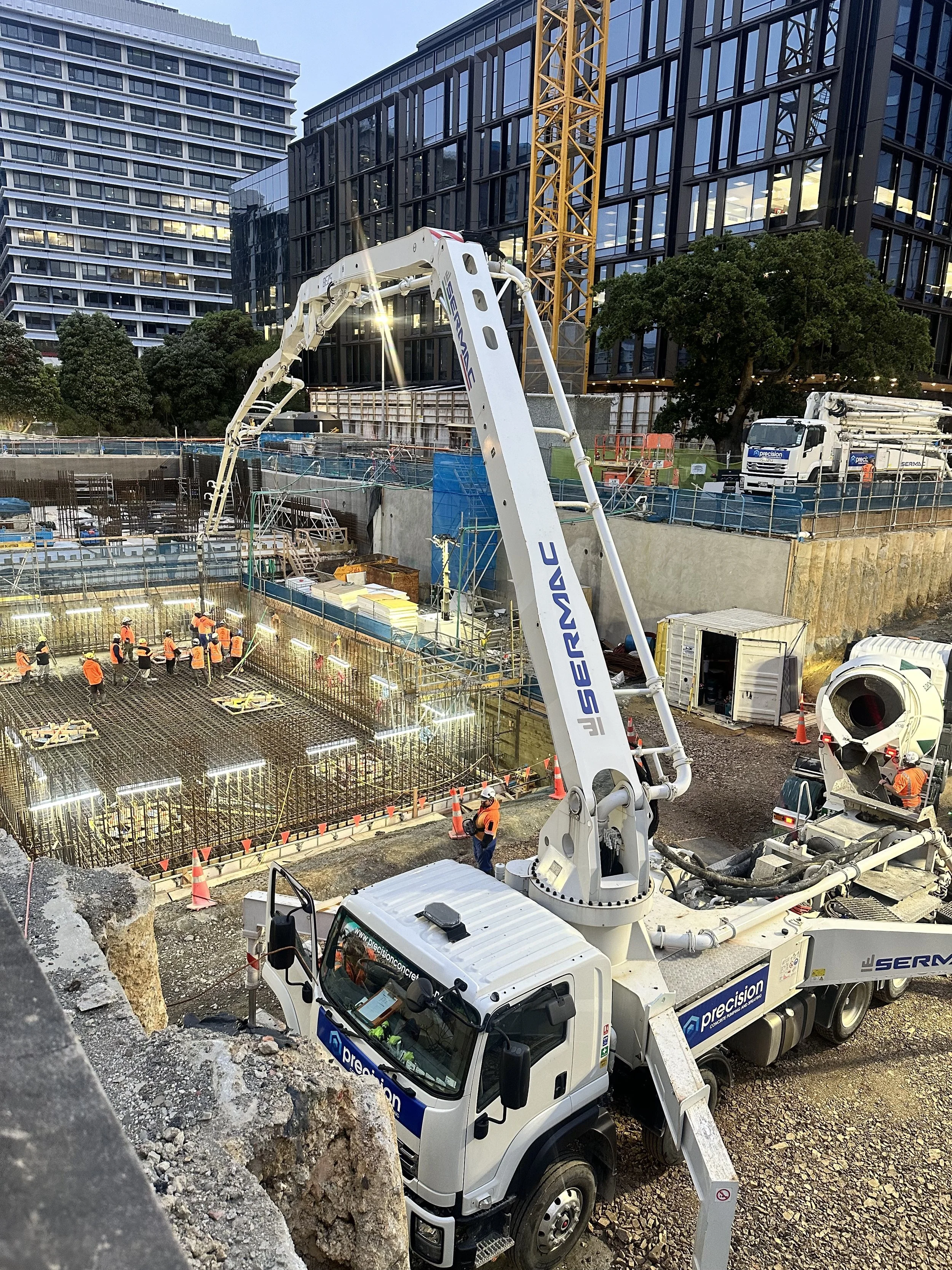 Construction site in an urban area with workers in orange safety vests, concrete reinforcement rebar, a large crane, and construction trucks, with modern glass office buildings in the background.