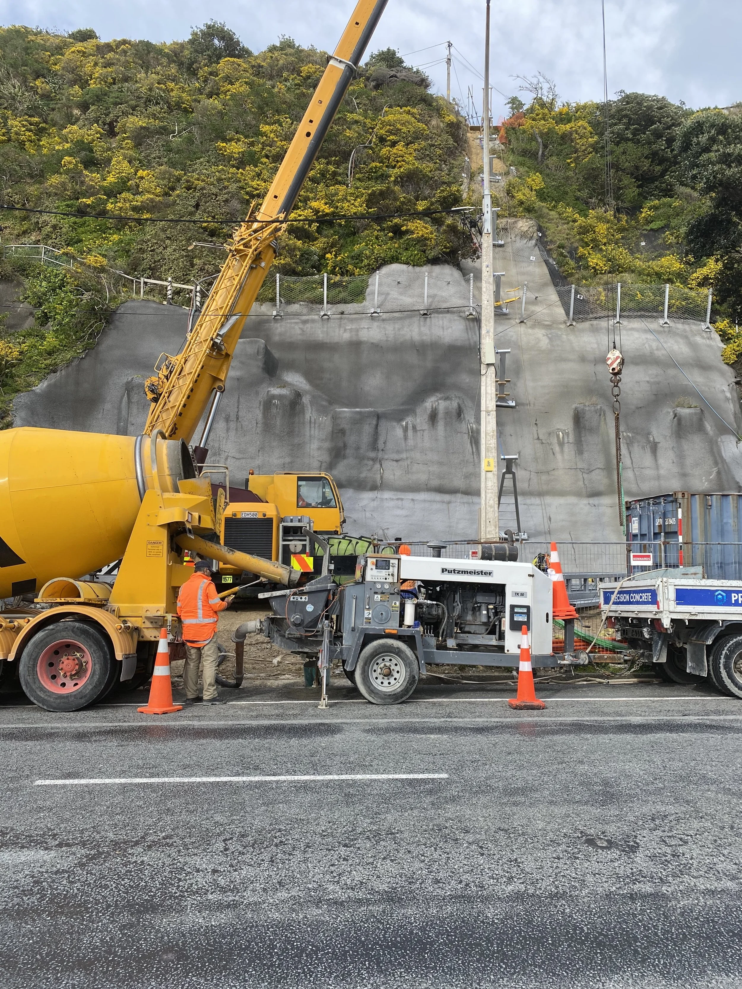 Construction workers and equipment installing or repairing a concrete retaining wall on a hillside, with orange cones marking the work area and a backdrop of green hillside and cloudy sky.