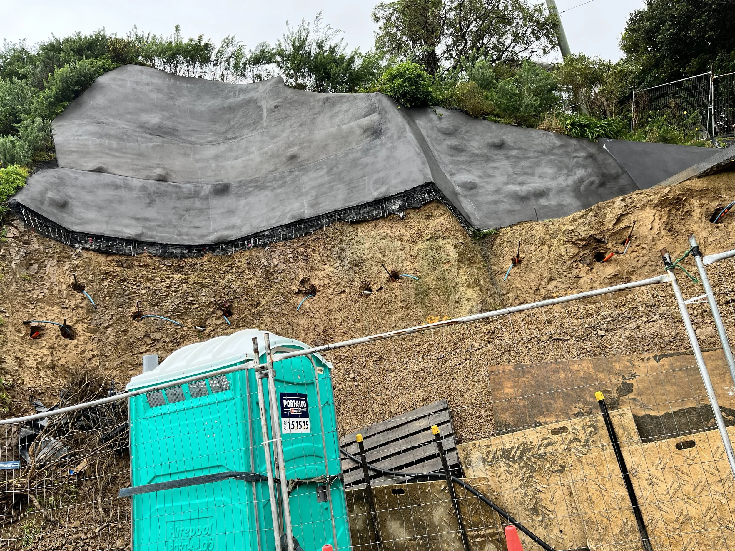 Construction site with exposed earth, a black waterproof membrane on a slope, and a portable toilet near a safety fence.