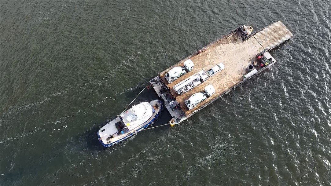 An aerial view of a boat transporting cars on a waterway, with the boats attached to a flat barge carrying several additional vehicles.