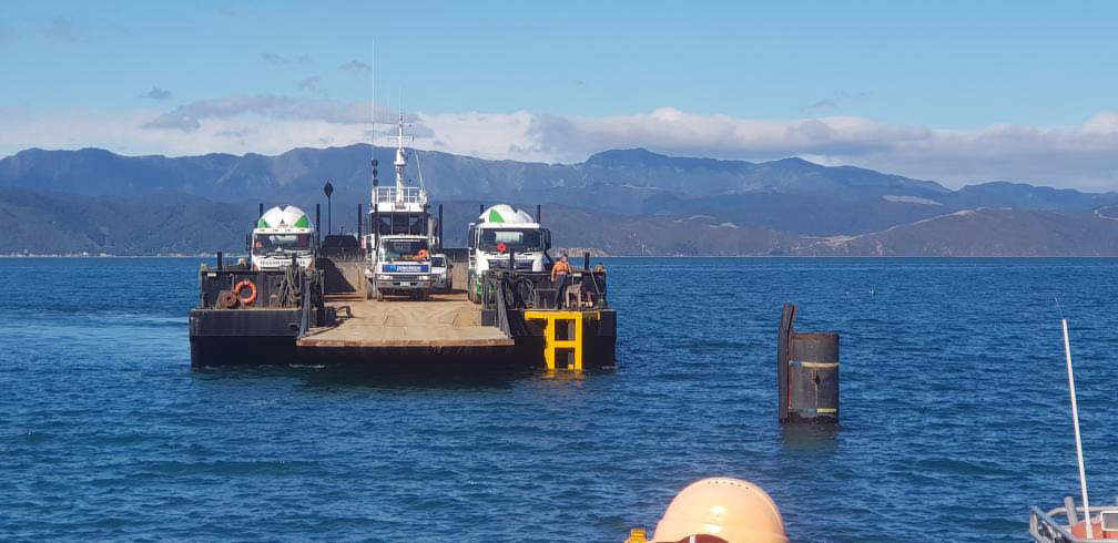 Ferry boat with trucks and vehicles on a calm body of water, with mountains in the background.