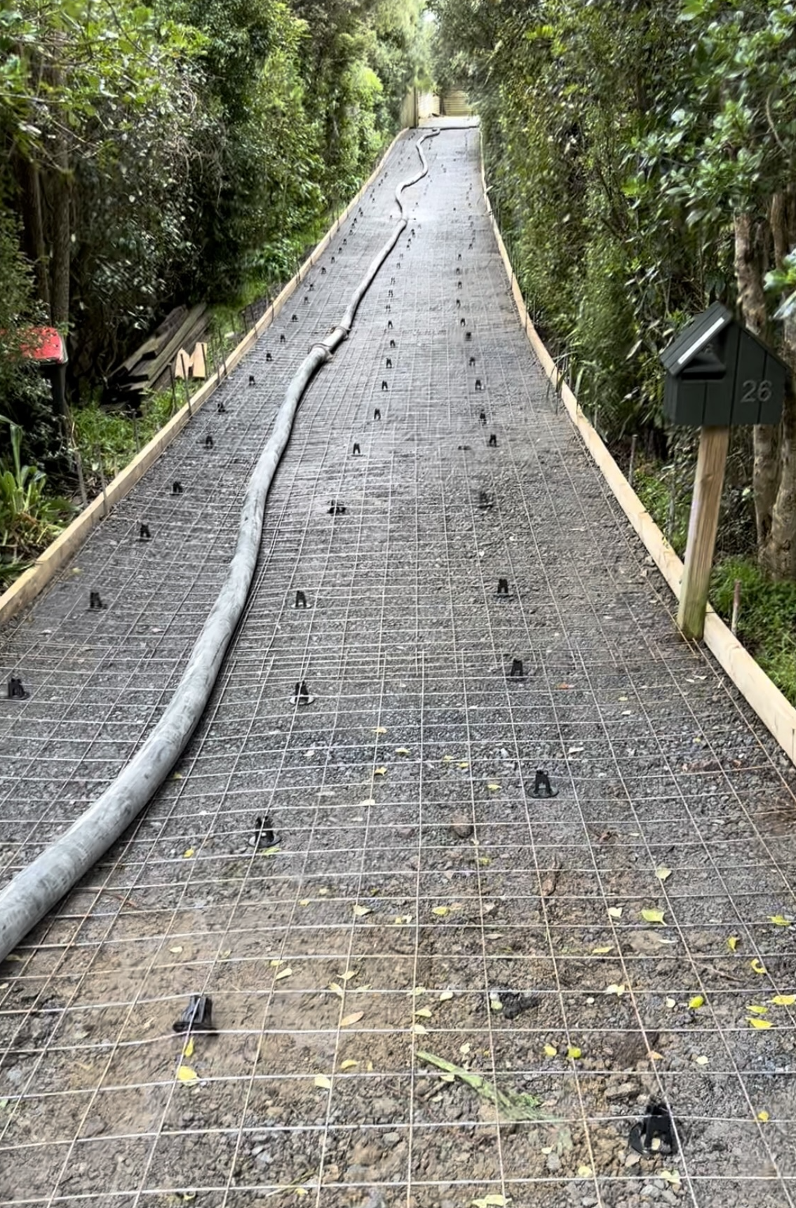 A pathway under construction with a metal grid surface, surrounded by lush green bushes and trees, with a hose lying down on the pathway.