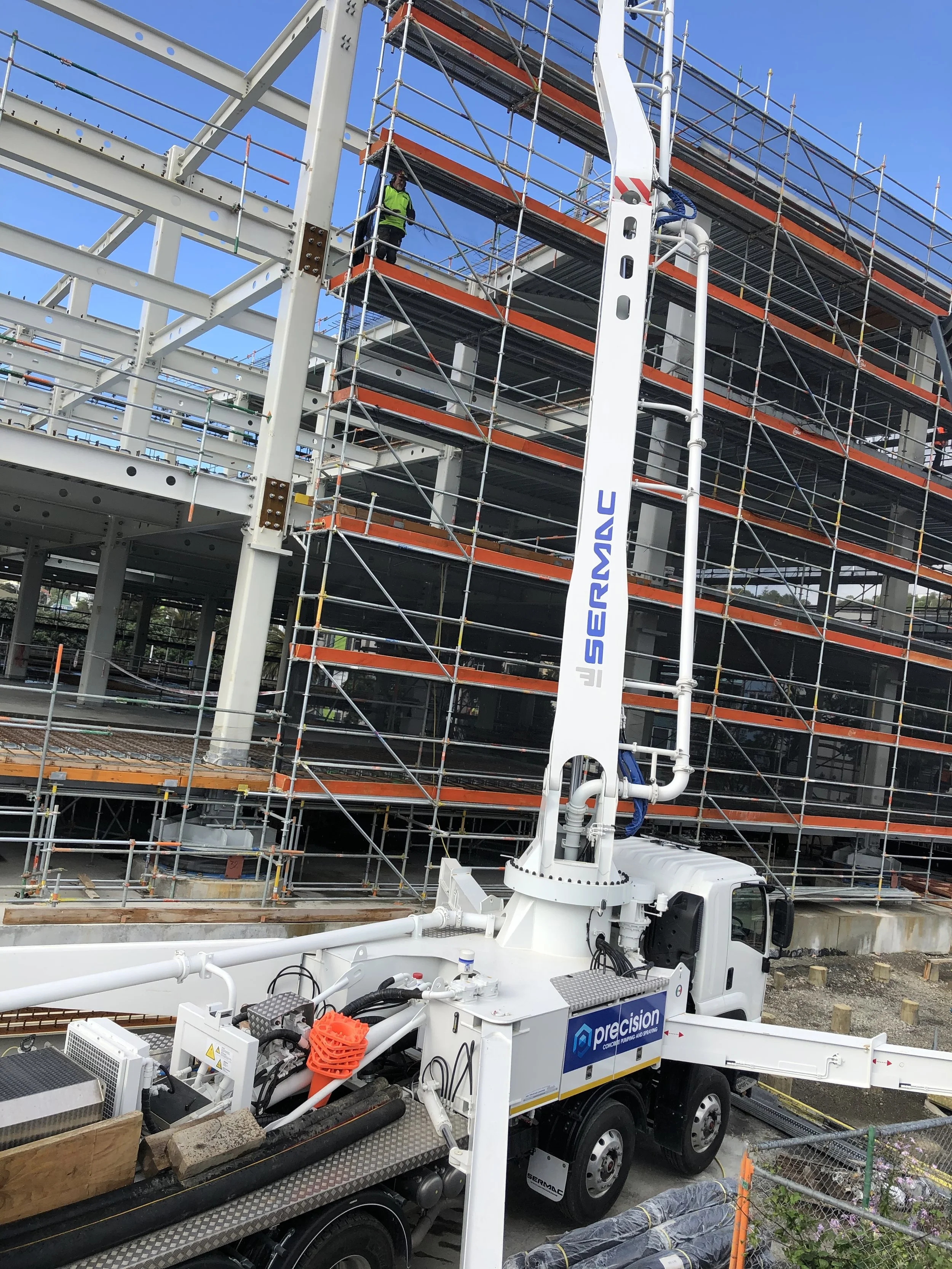 Construction site with a large white crane and scaffolding around a building structure. There is a worker in a green vest on the scaffolding. Construction materials and equipment are visible on the ground.