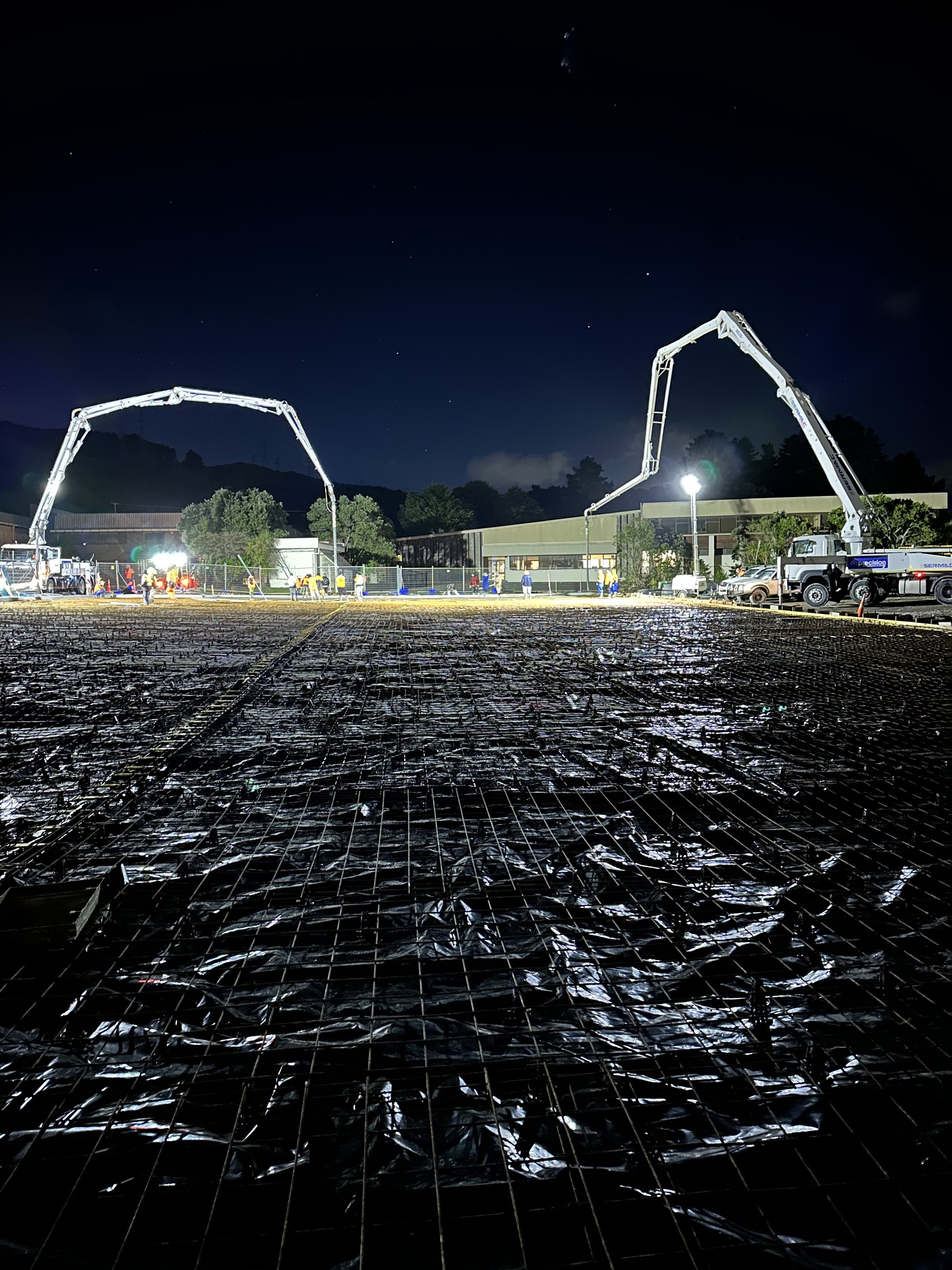 Nighttime construction site with workers, power trucks with cranes, illuminated by bright streetlights, with a building, trees, and a mountain in the background.