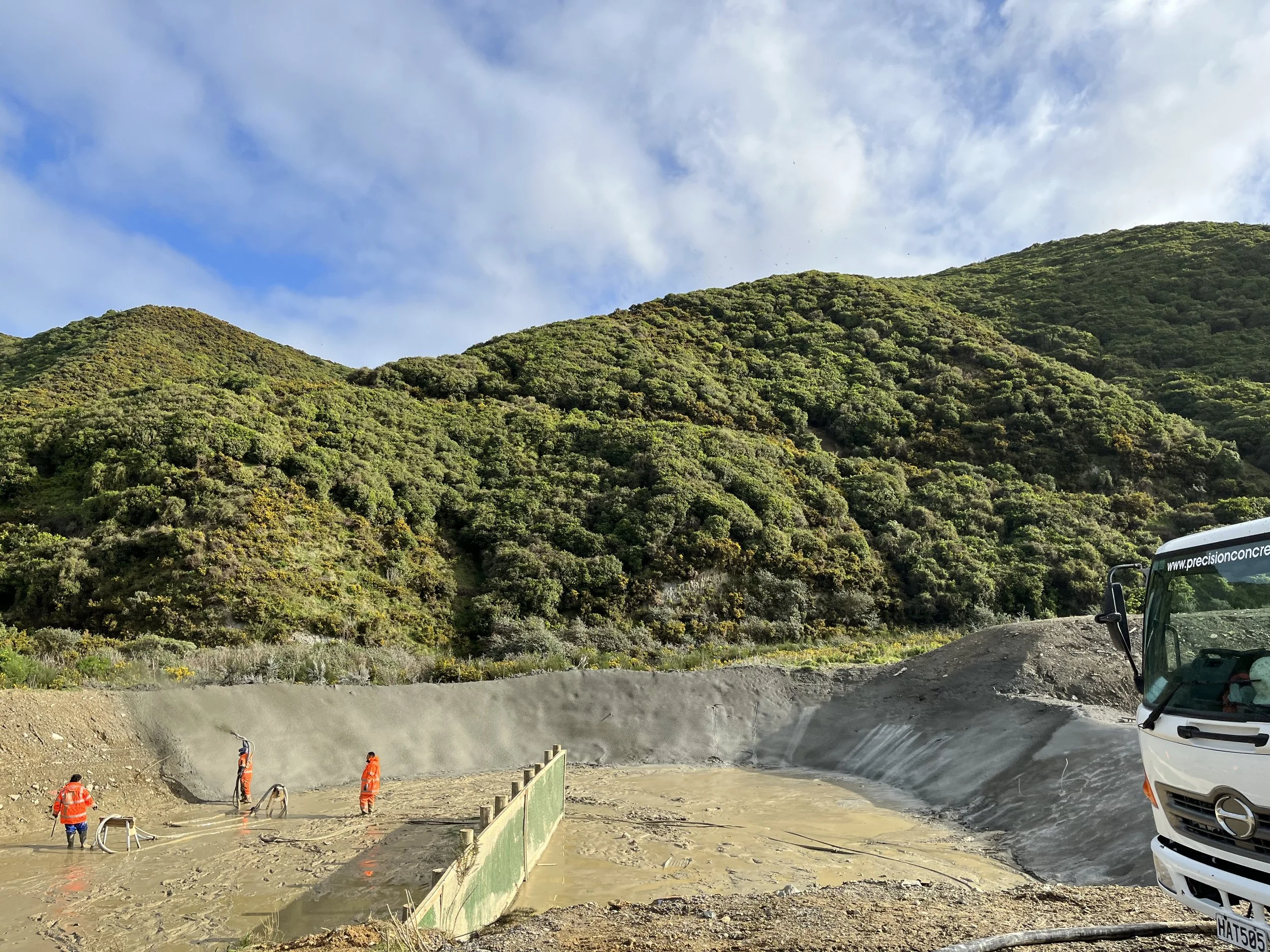 Construction workers in orange safety vests working on a concrete paving project at the base of green, rolling hills under a partly cloudy sky, with vehicles parked nearby.