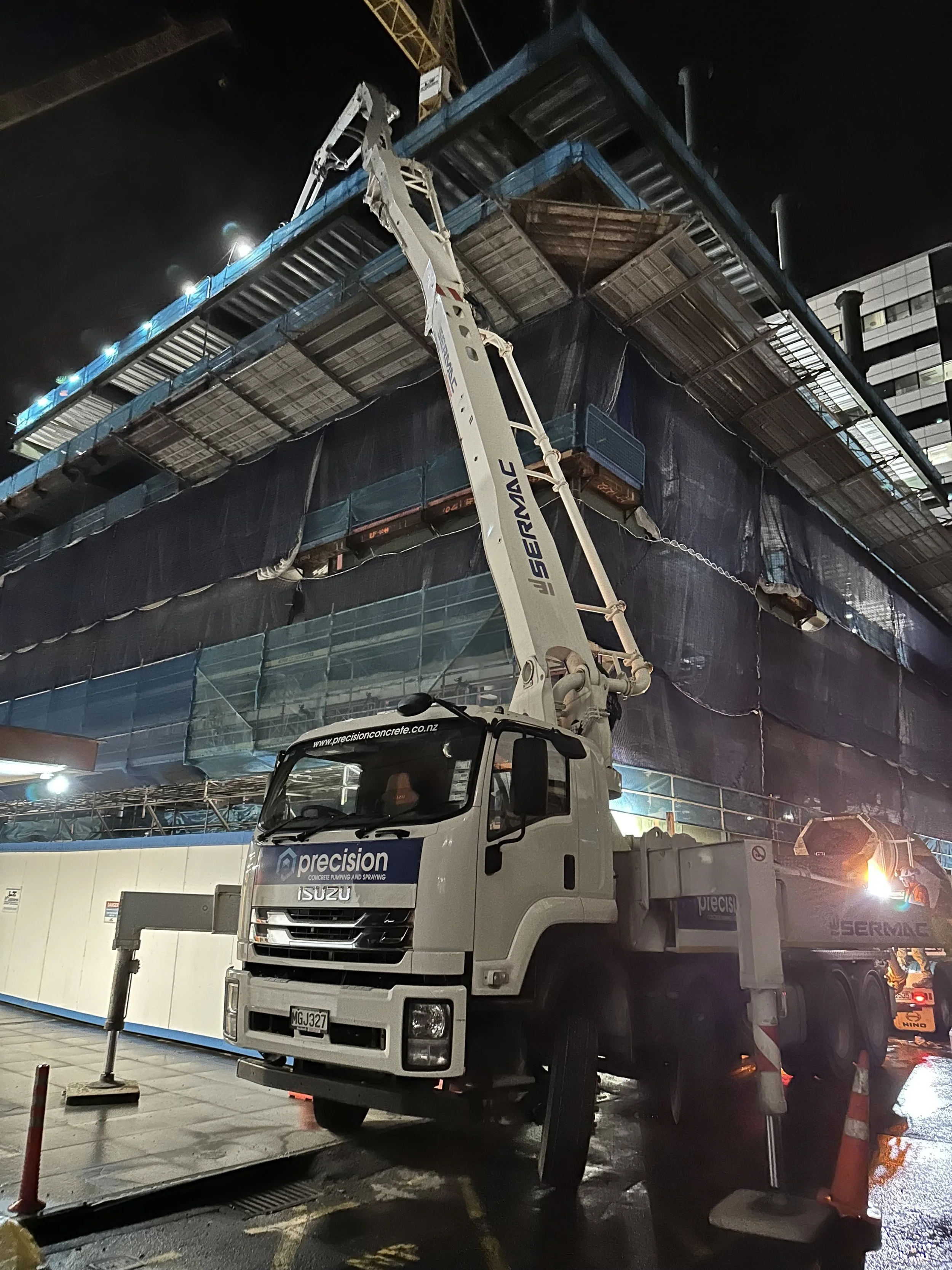A large construction truck with a crane arm is working at a building site at night. The truck has the branding 'precision' and 'ISUZU'. The building under construction has multiple floors with blue and black safety netting. There are construction cones and barriers on the ground, and the area is lit by bright artificial lighting.