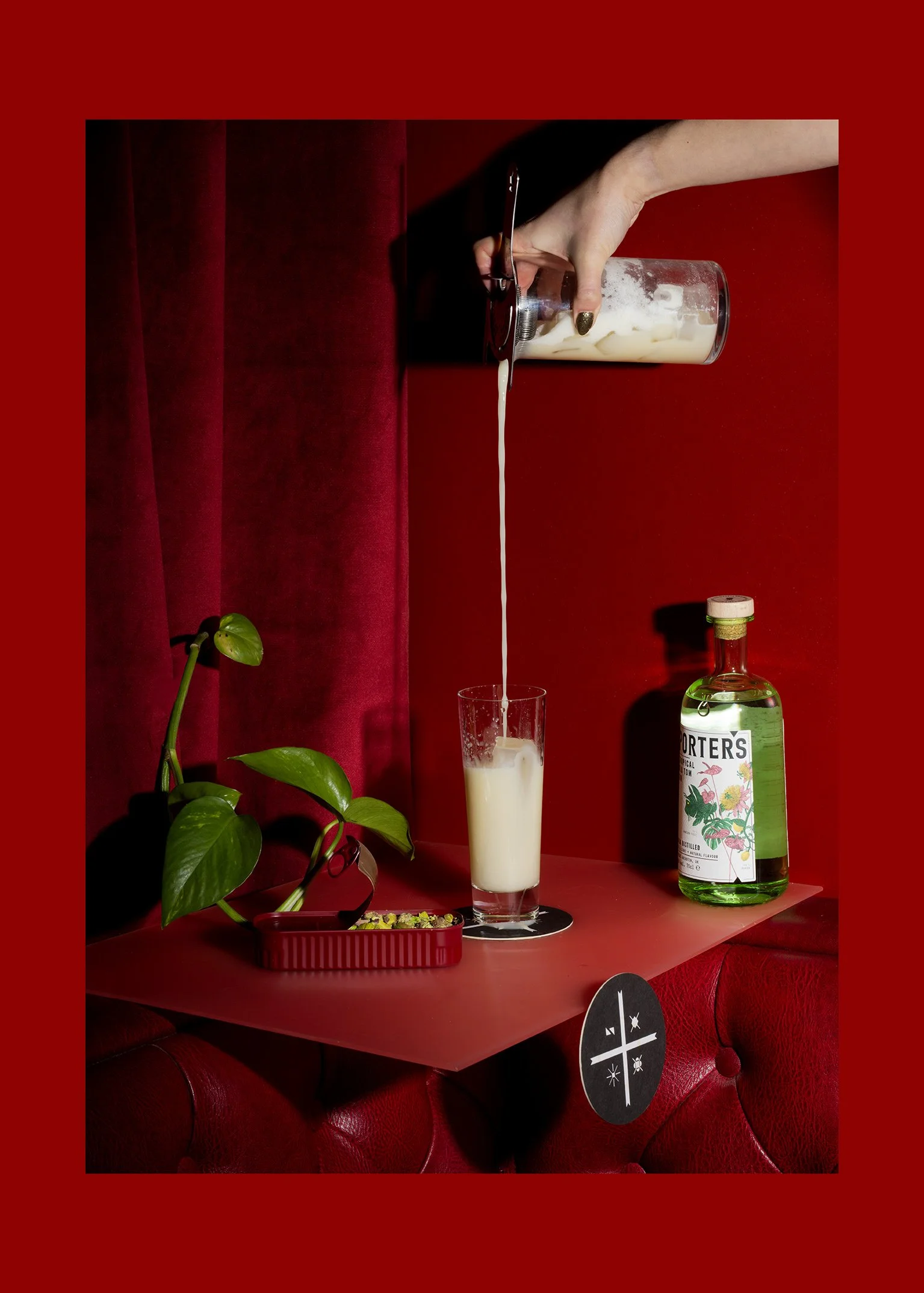 A hand pouring a creamy drink from a glass container into a tall glass, with a green plant and a bottle of porters tonic on a red table against a red background.