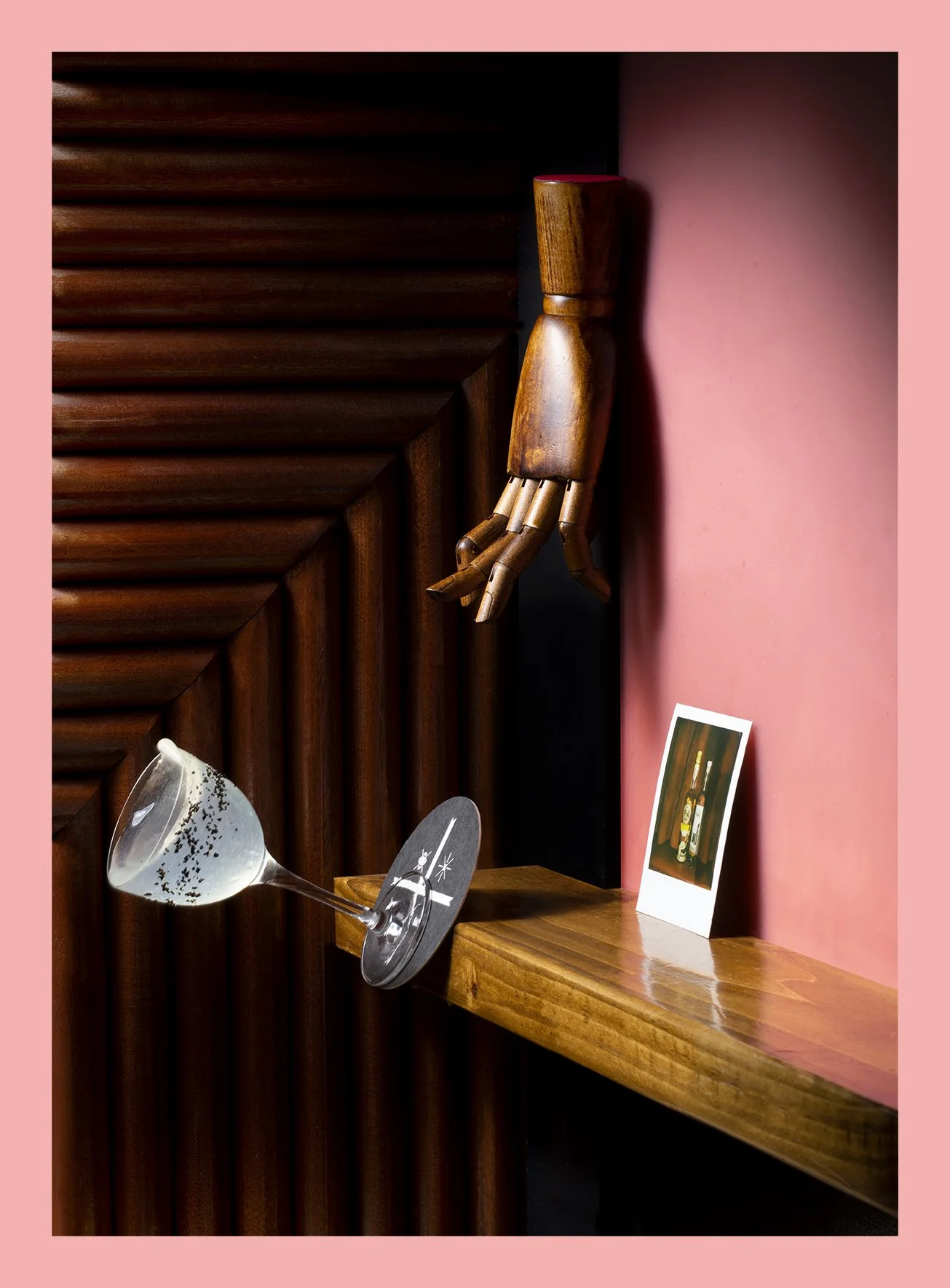 A wooden hand sculpture mounted on a brown wood-paneled wall, a tilted martini glass with black specks on a wooden shelf, and a small picture of liquor bottles leaning against a pink wall.
