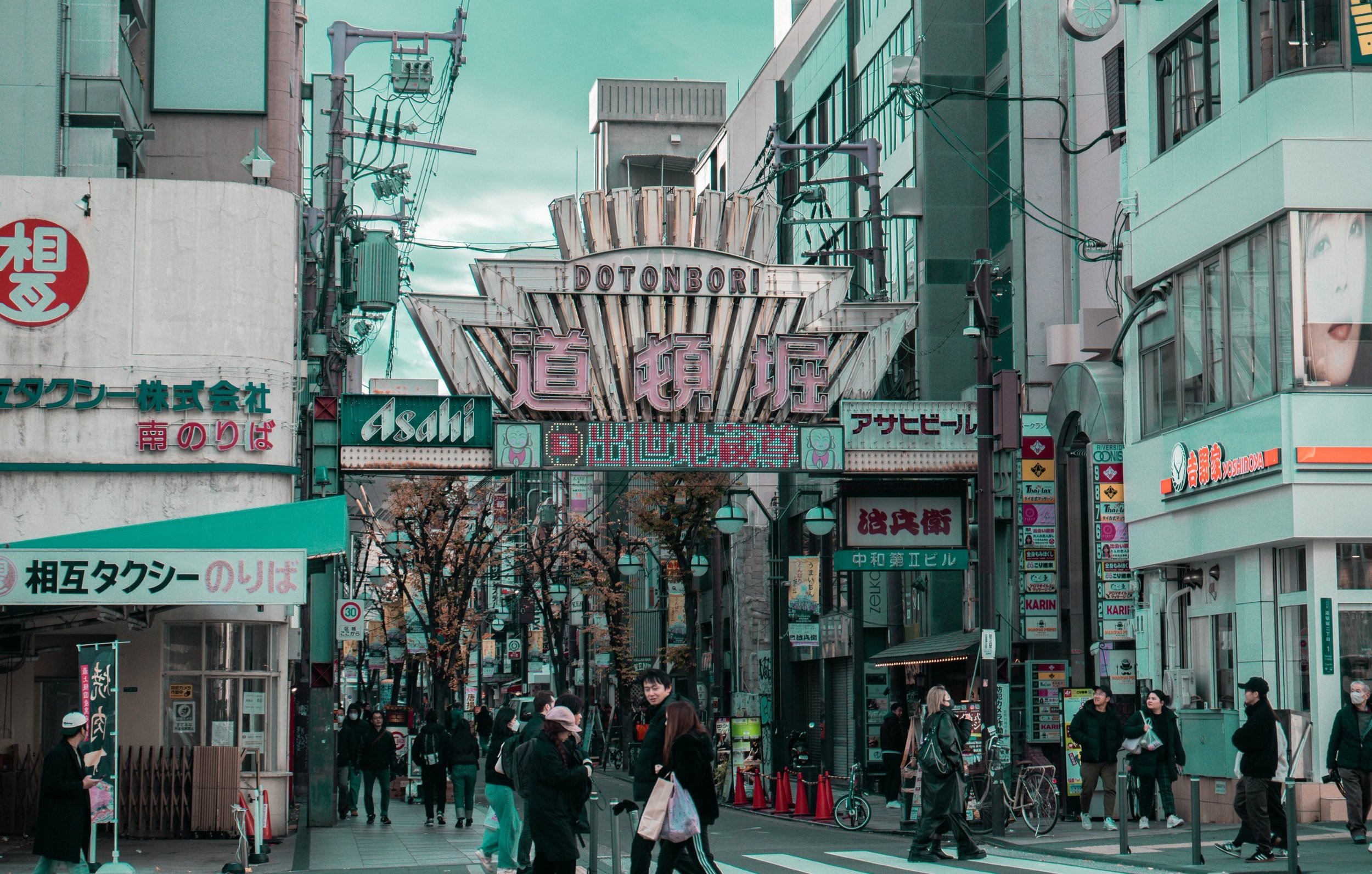 Urban street scene in Japan with signs in Japanese, pedestrians walking, bicycles parked, and storefronts along the street.