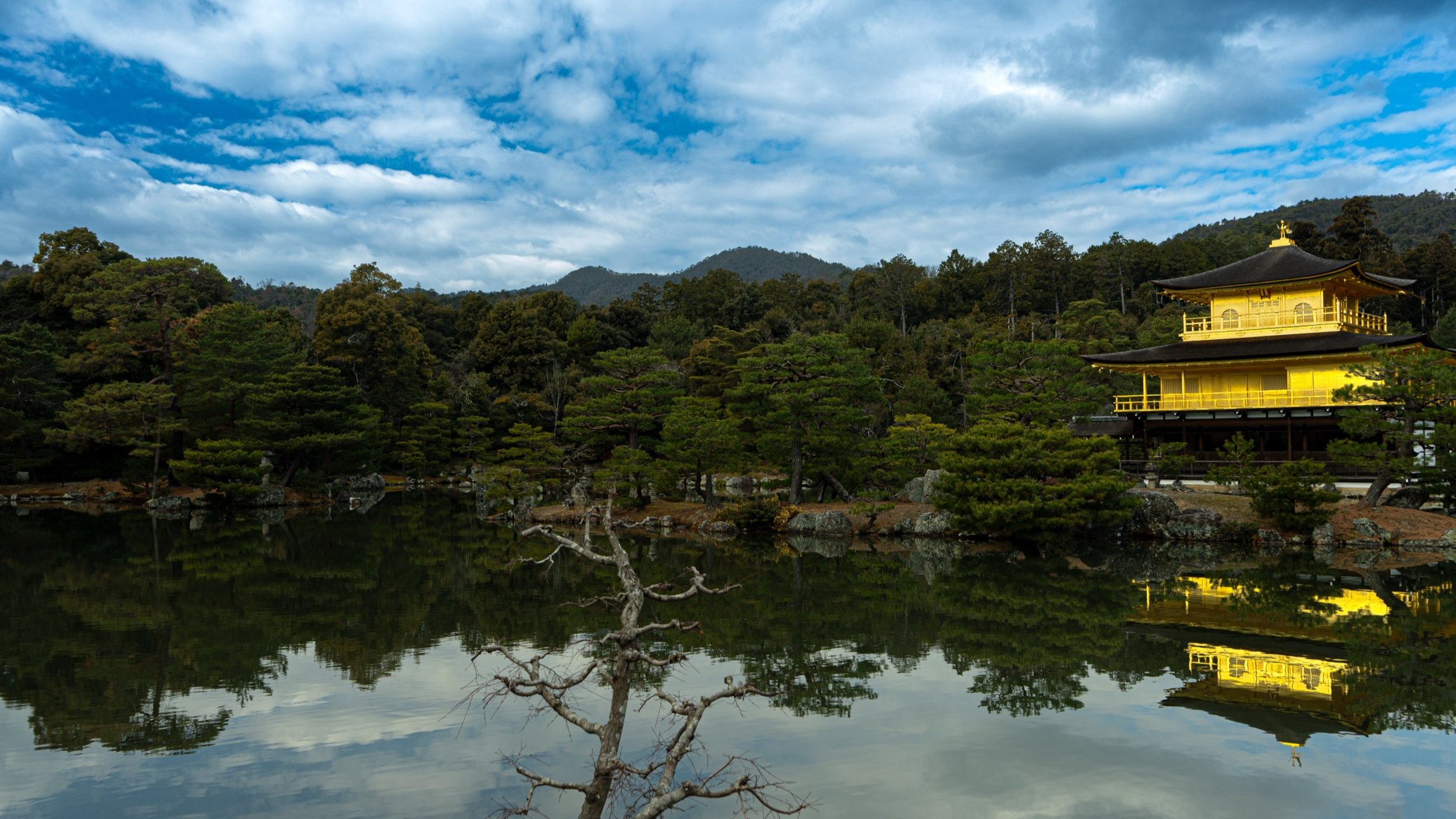 A traditional Japanese temple with yellow walls on the water's edge, surrounded by lush green trees and mountains, with a partly cloudy sky reflecting in the lake.