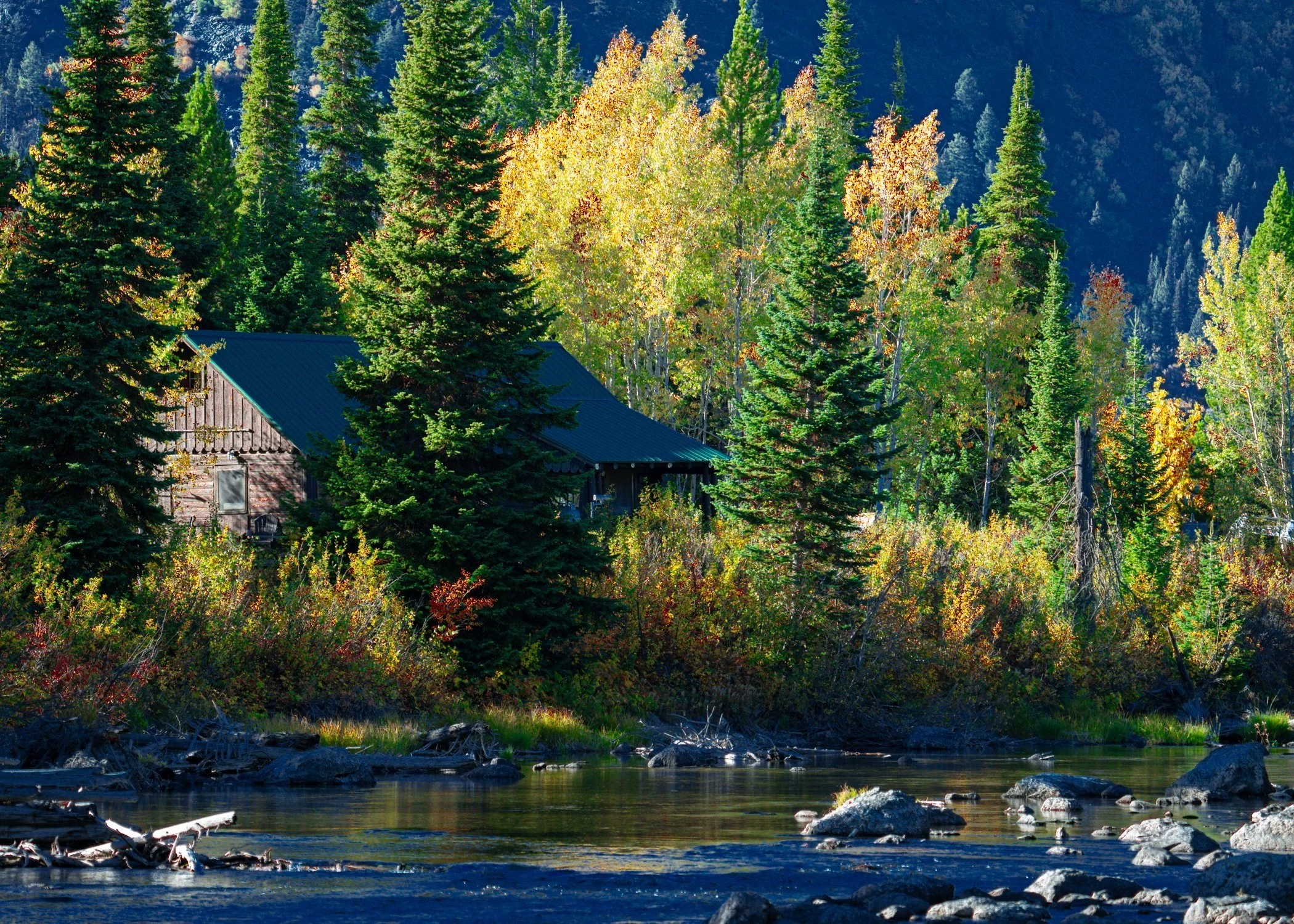 A wooden cabin with a blue roof surrounded by evergreen trees, colorful autumn foliage, and a river in the foreground, in a mountain forest setting.