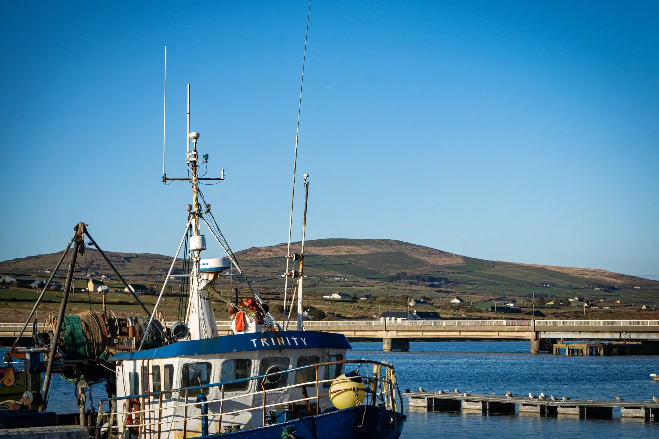 A fishing boat named Trinity docked at a harbor with a bridge and rolling hills in the background under a clear blue sky.