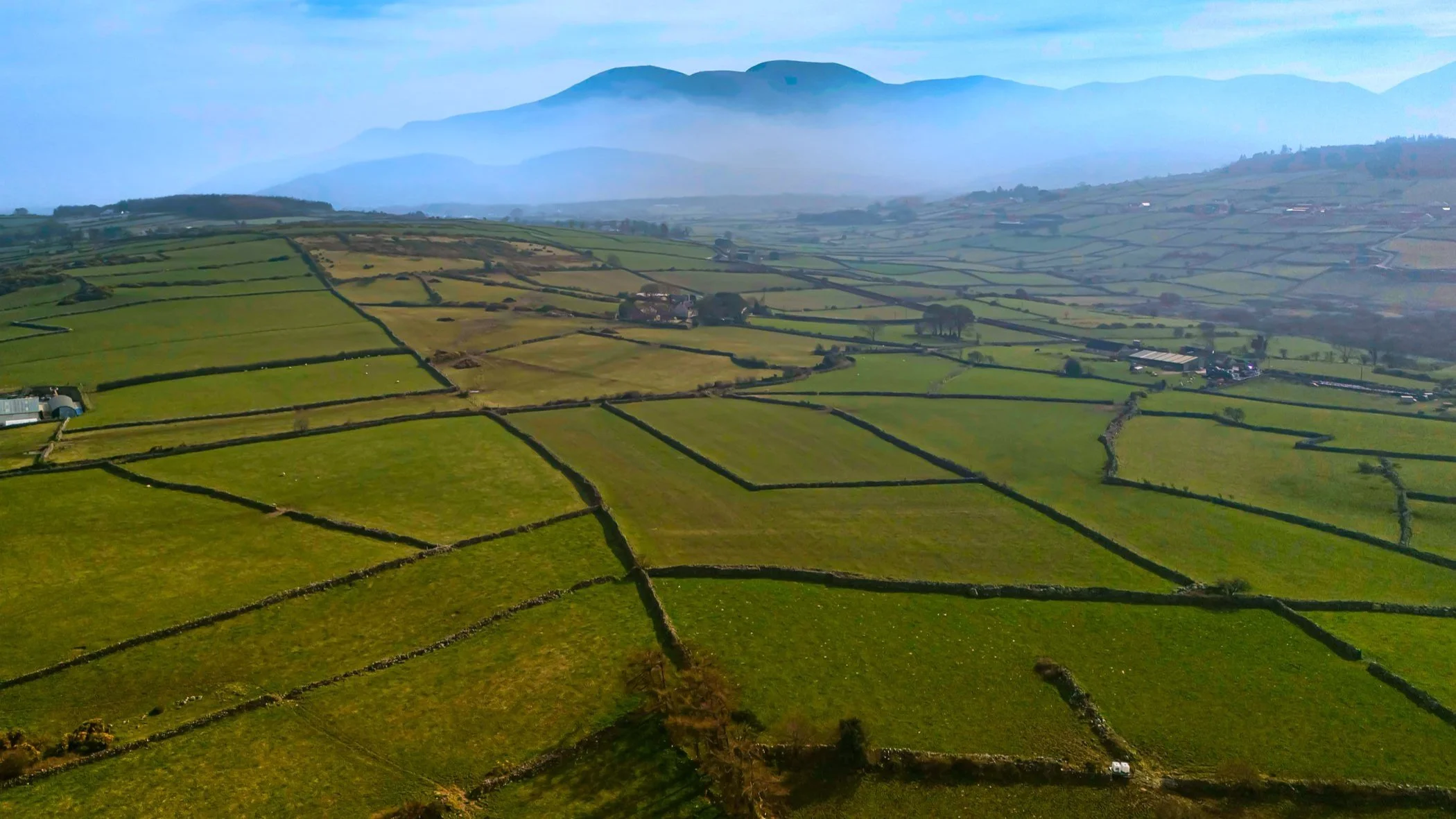 Aerial view of green agricultural fields separated by stone walls, with a distant mountain range under a partly cloudy sky.
