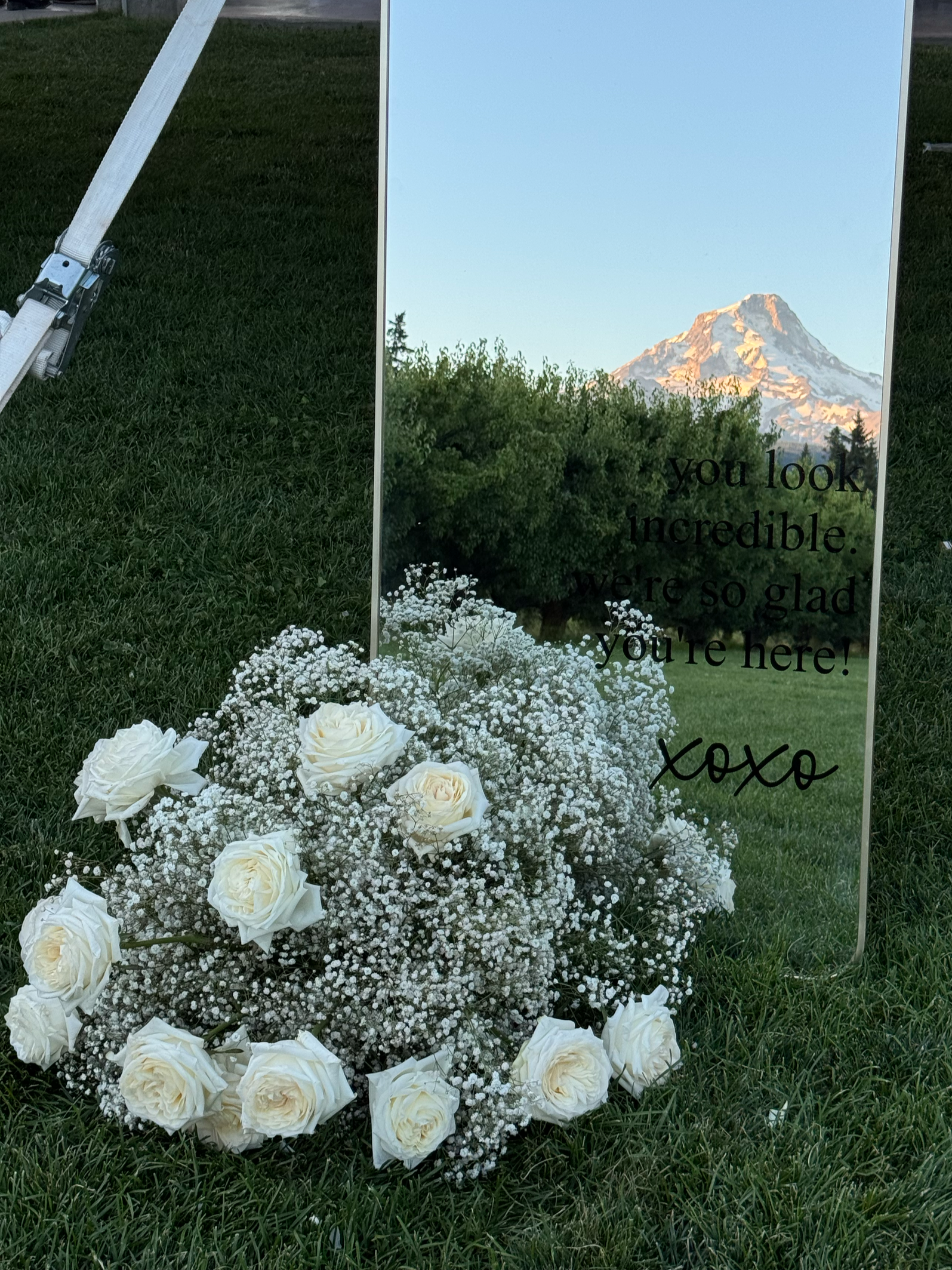 A mirror with an inscription and a bouquet of white roses and baby's breath on green grass, with Mount Hood in the background.