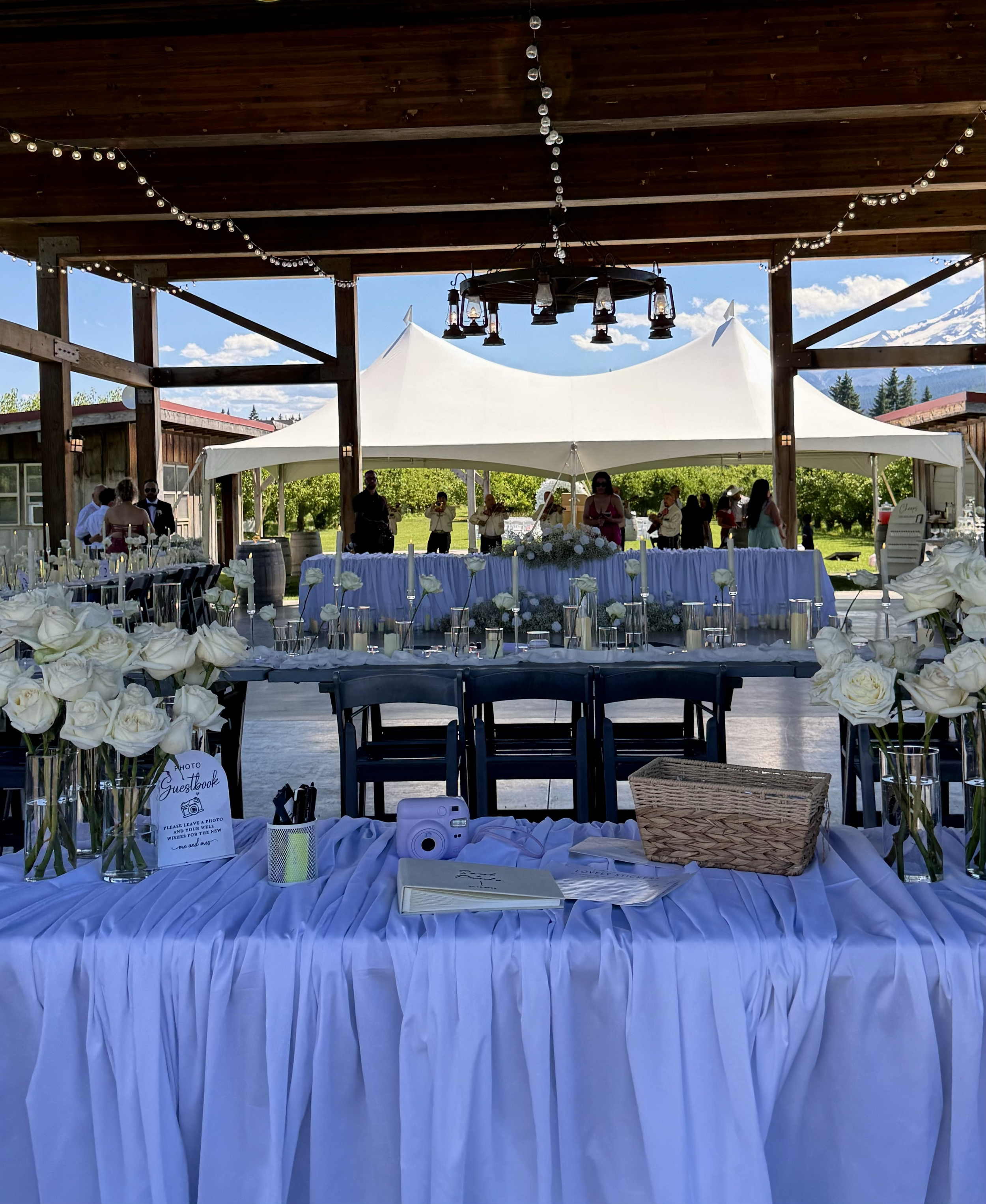 Outdoor wedding reception setup with long tables adorned with white roses, candles, and a guestbook, under a wooden pavilion with string lights, and a large tent in the background, with mountains and trees in the distance on a sunny day.