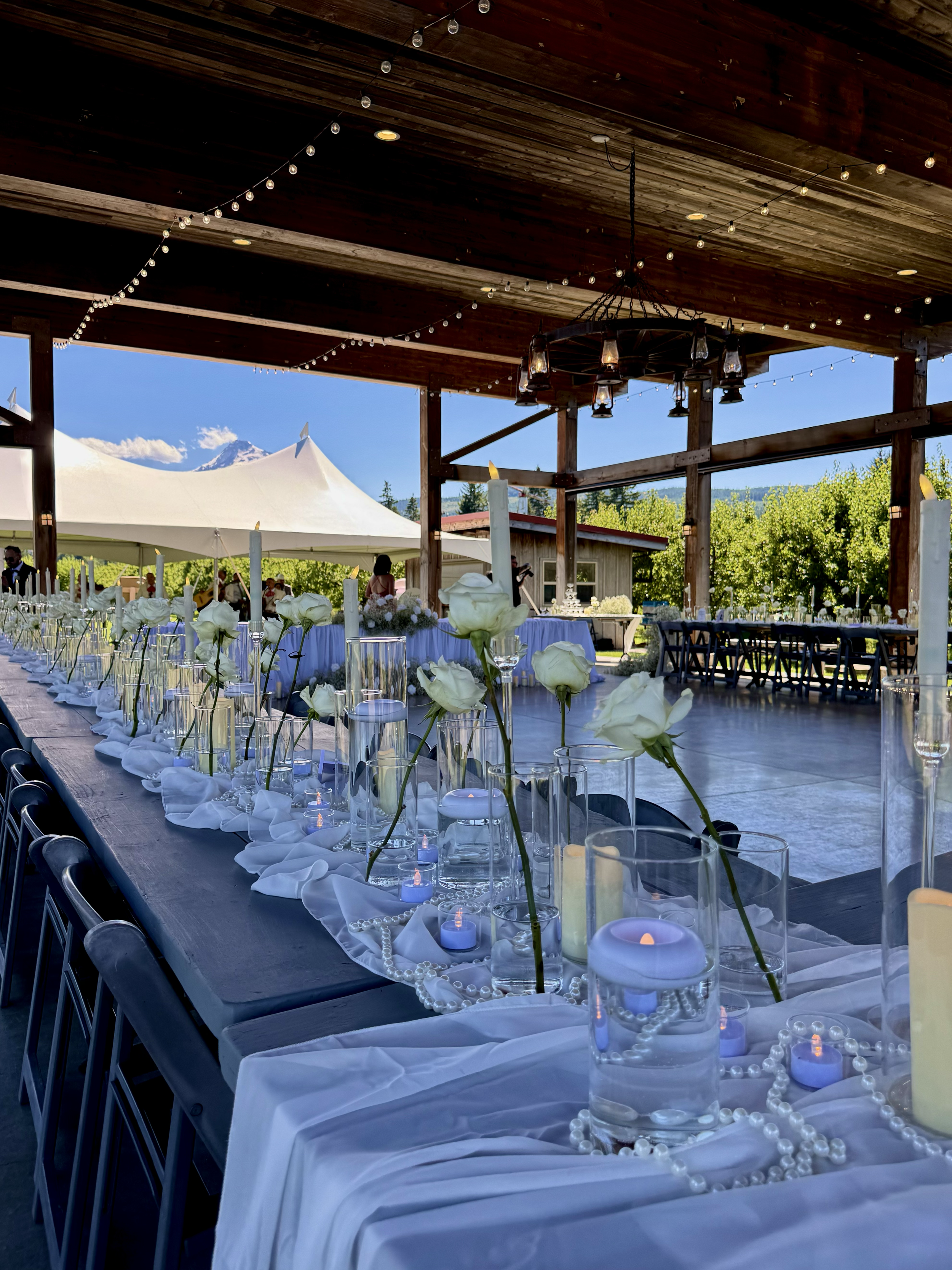 Decorated outdoor wedding reception table under a wooden pavilion with white roses, candles, pearls, and string lights, overlooking a tent and mountain in the background.