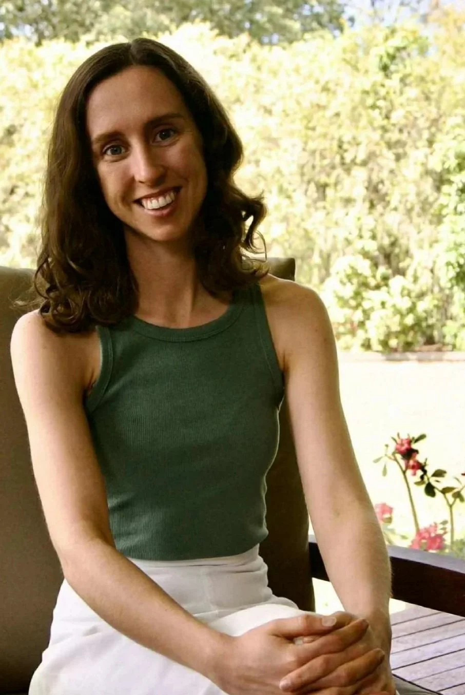 A woman with brown, curly hair smiling and sitting outdoors, wearing a sleeveless green top and a white skirt, with greenery and pink flowers in the background.