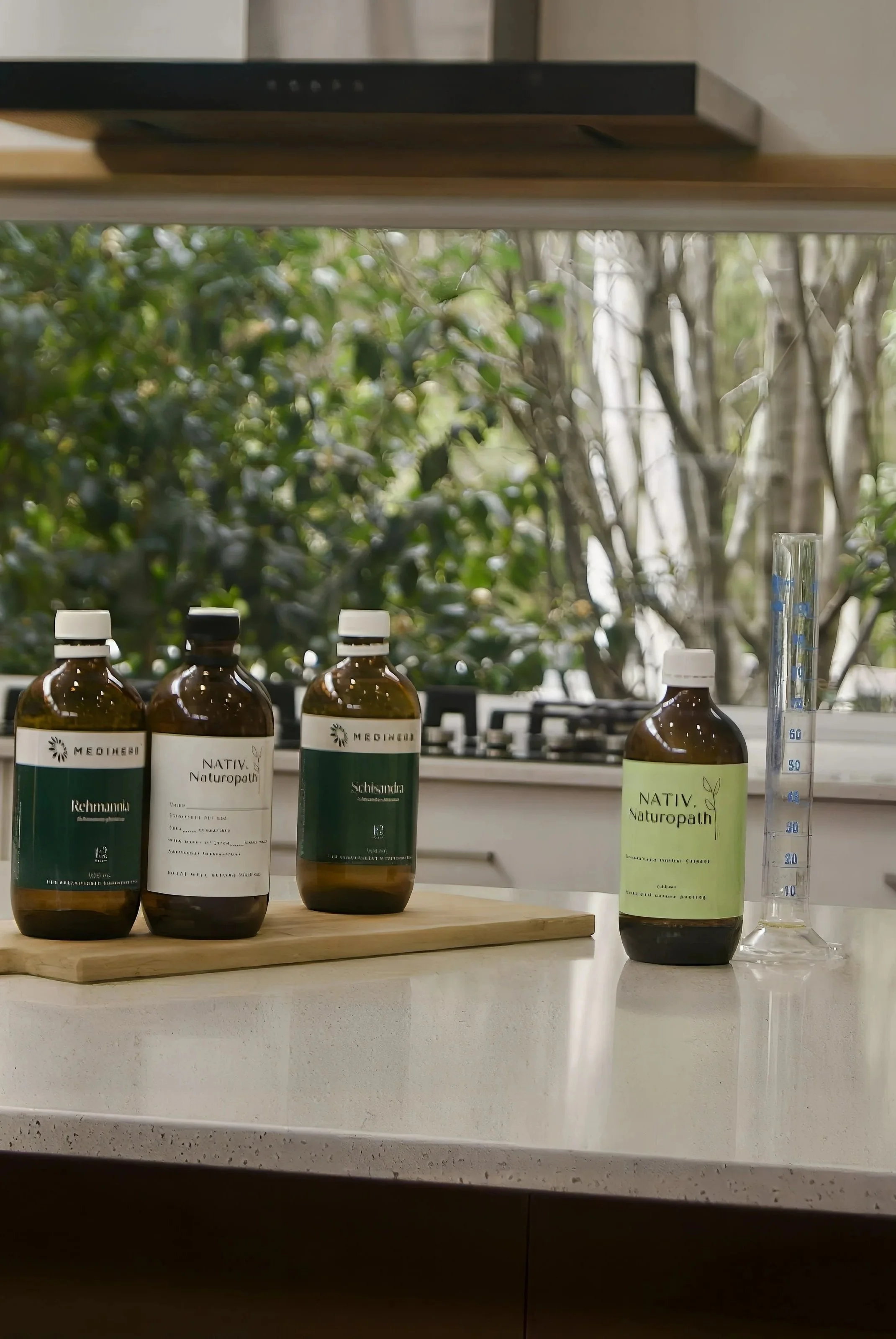 Four medicine bottles with labels and a measuring tube on a kitchen counter, with a window showing trees in the background.