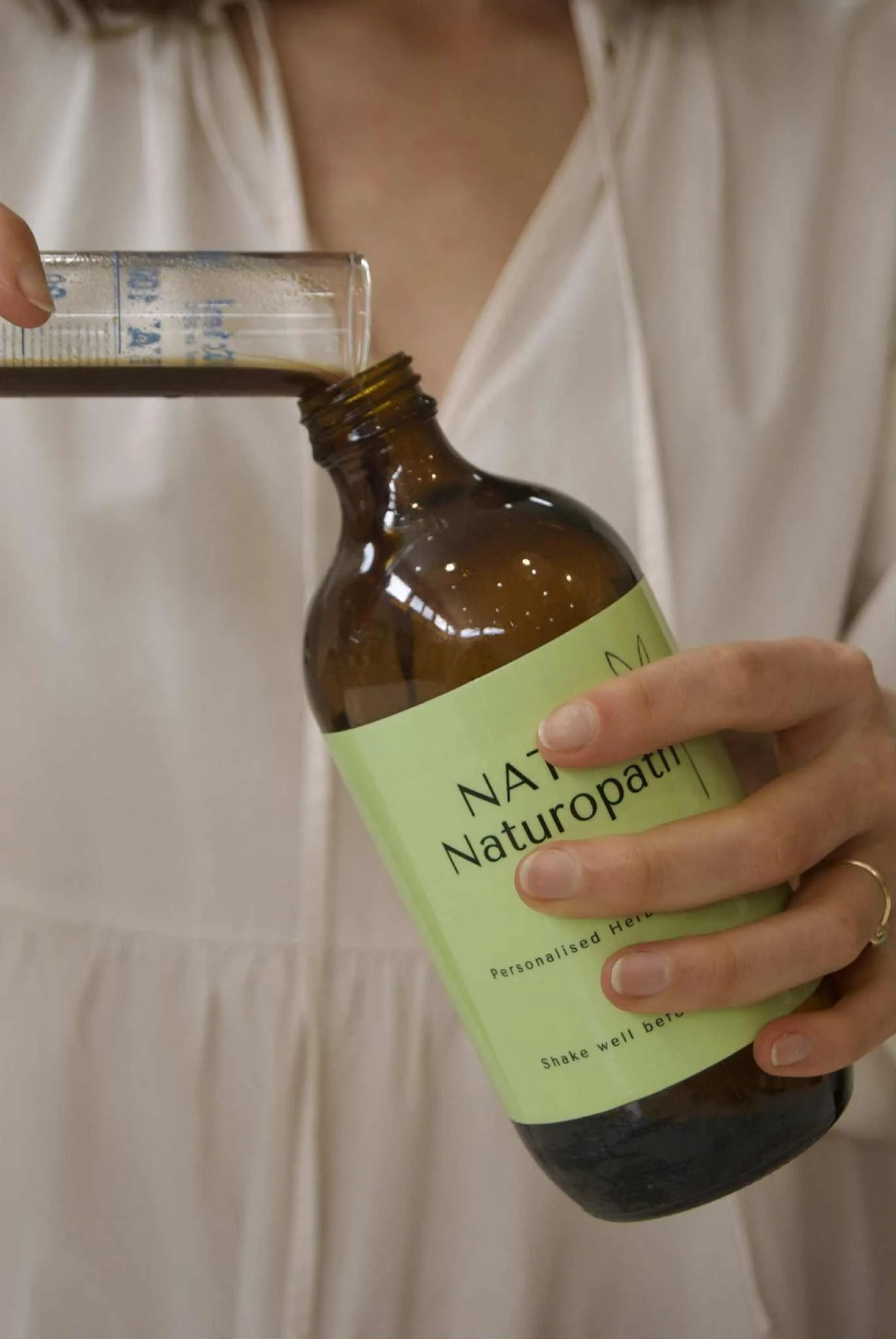 Person pouring herbal liquid from a small beaker into a brown bottle labeled 'NATUROPATHY'.