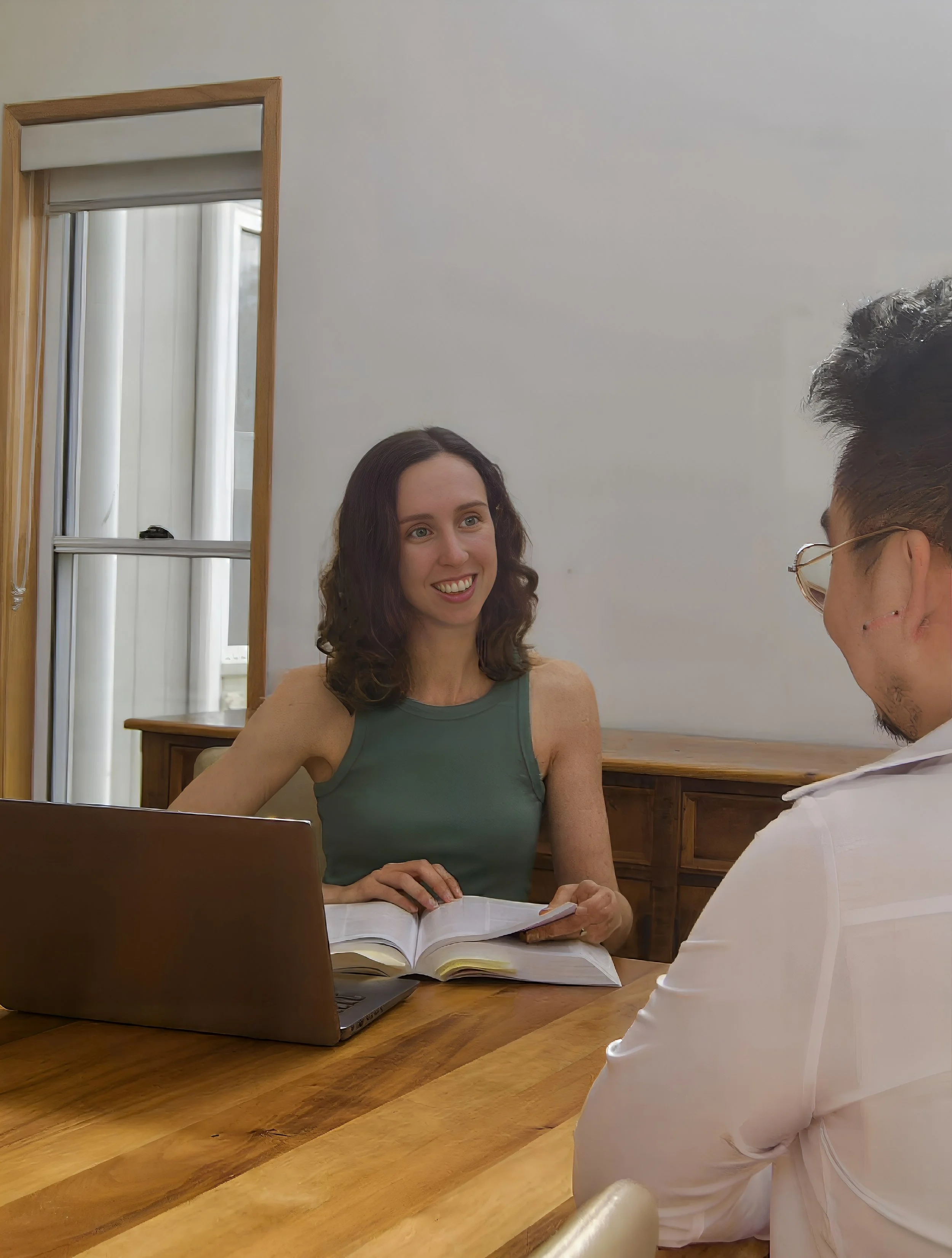 A woman and a man having a conversation at a wooden table, with a laptop open and a book in front of her, in a room with a window and wooden furniture.