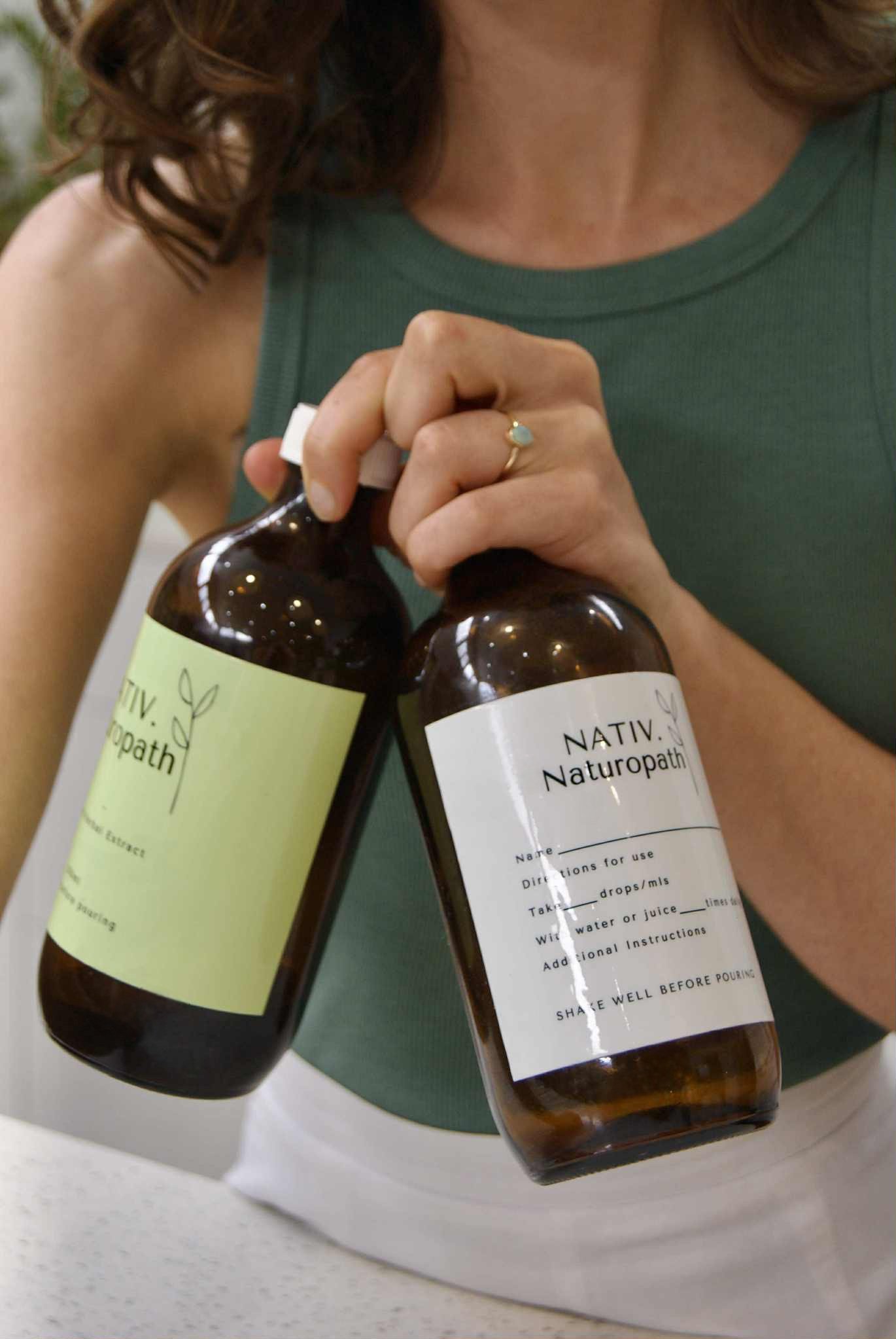 A woman holding two brown glass bottles labeled 'NATIV. Naturopath,' one with a green label and one with a white label, next to her chest. The woman is wearing a green tank top and has a ring with an opal on her finger.