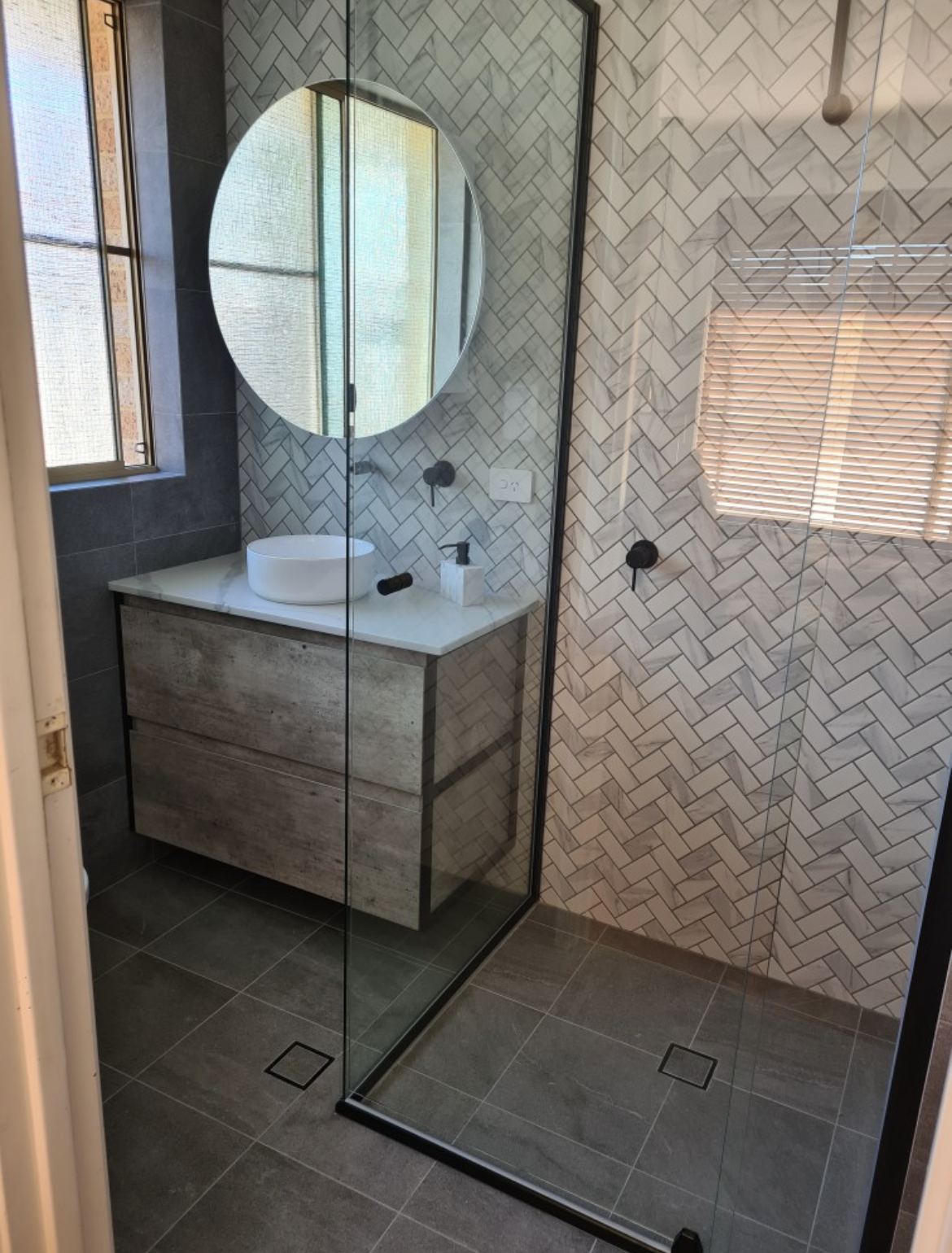 Modern bathroom with a walk-in glass shower, herringbone patterned tile on the walls, a round mirror, a vessel sink on a vanity, and a window with wooden blinds.