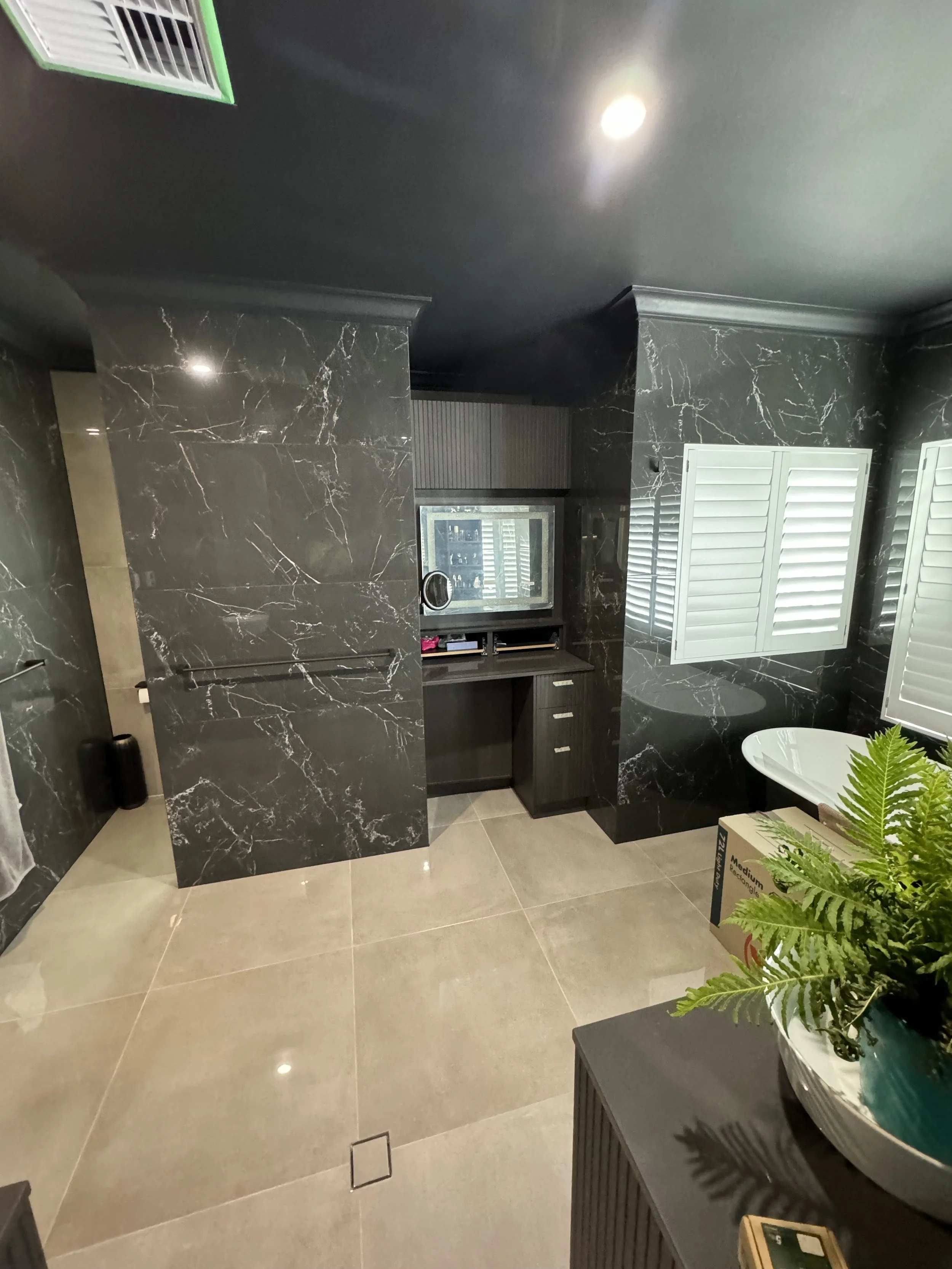 Bathroom with black marble walls, beige tiled floor, window with white shutters, and a potted plant on a table.