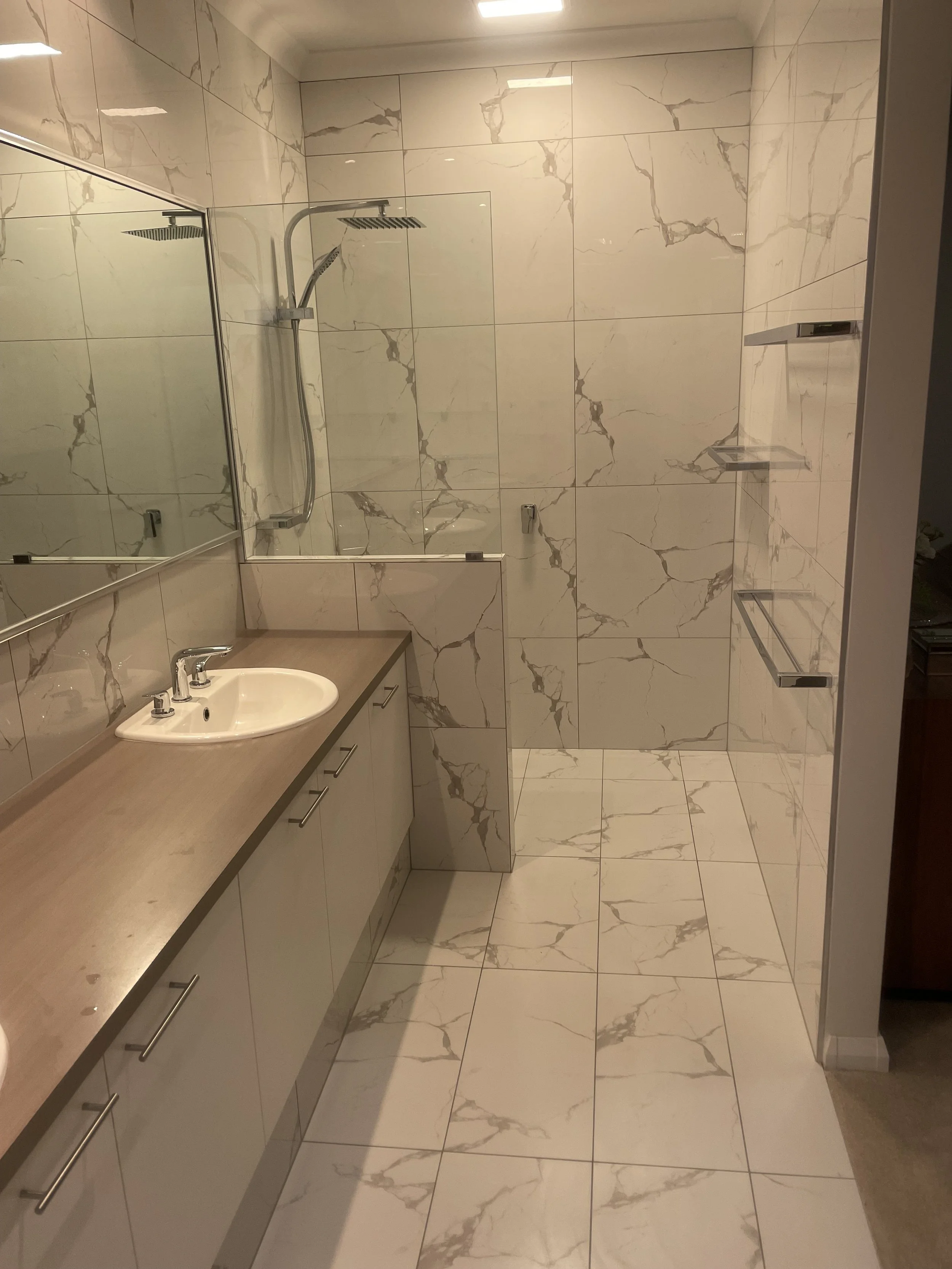 Bathroom with marble tiles, a vanity with a sink, a large mirror, a walk-in shower with glass door, and built-in shelves in the shower area.