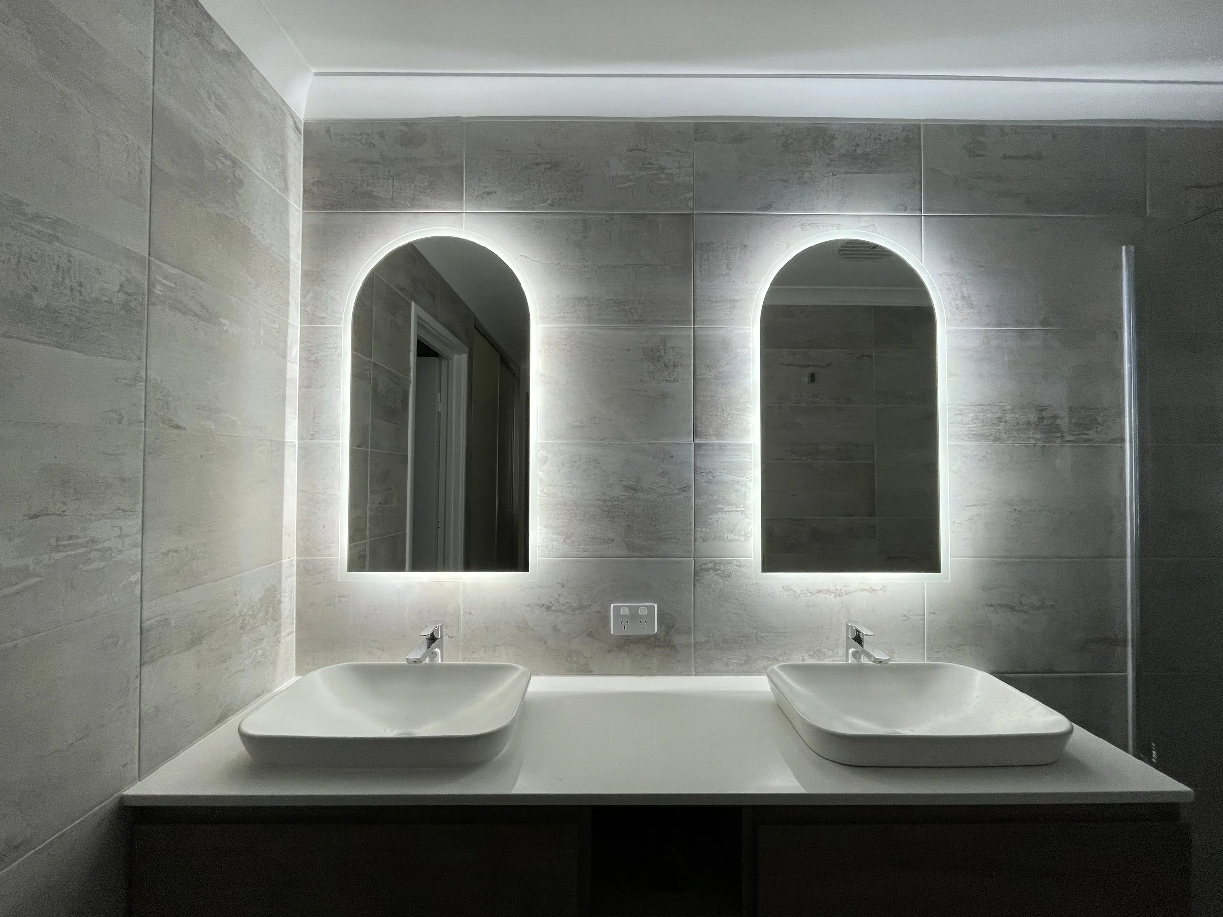 Modern bathroom with two rectangular vessel sinks on a white countertop, illuminated mirrors with LED backlighting, and a wall socket between the sinks, all set against light gray tiled walls.