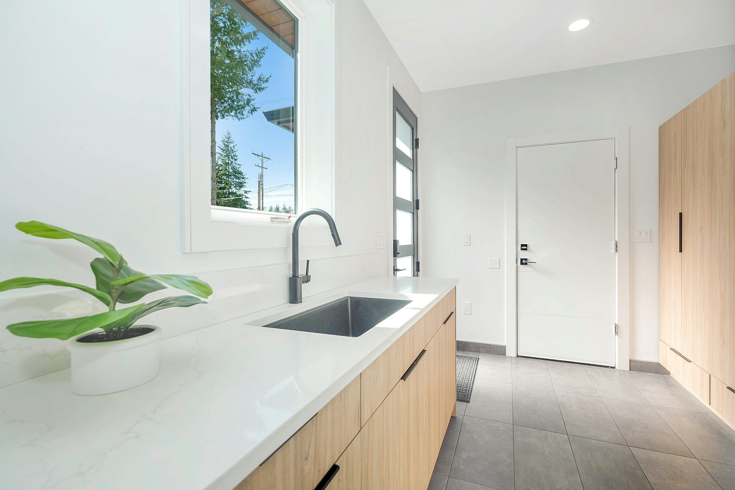 Modern kitchen with white walls, wooden cabinets, and a white countertop featuring a small potted plant. A black sink and gray faucet are installed near a window that shows outdoor trees and power lines. A door and a tall wooden cabinet are visible in the background.