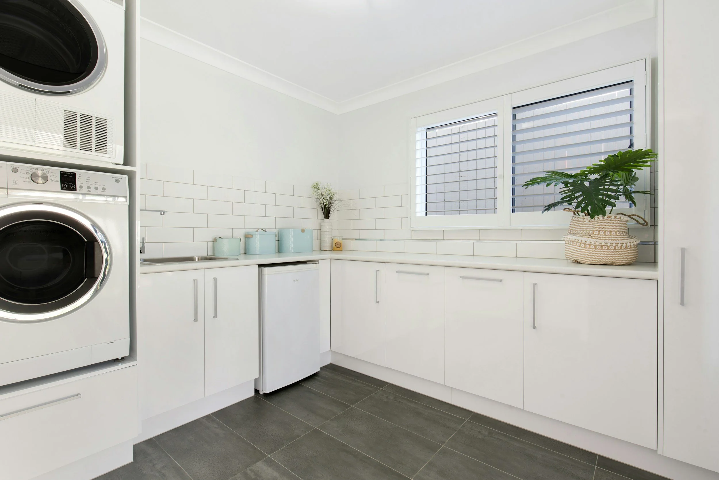 Modern white laundry room with front-loading washer and dryer, small refrigerator, and white cabinetry, adorned with a potted plant and decorative containers, illuminated by natural light from a window.