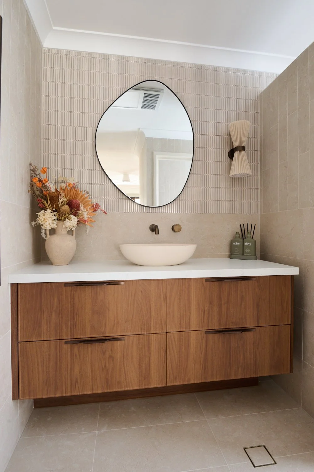Modern bathroom vanity with a round white vessel sink, black matte faucet, and a large oval mirror. Decor includes a vase of dried flowers on the left and green apothecary bottles with black reed diffusers on the right. Wood cabinet with four drawers, set against beige tiled walls.