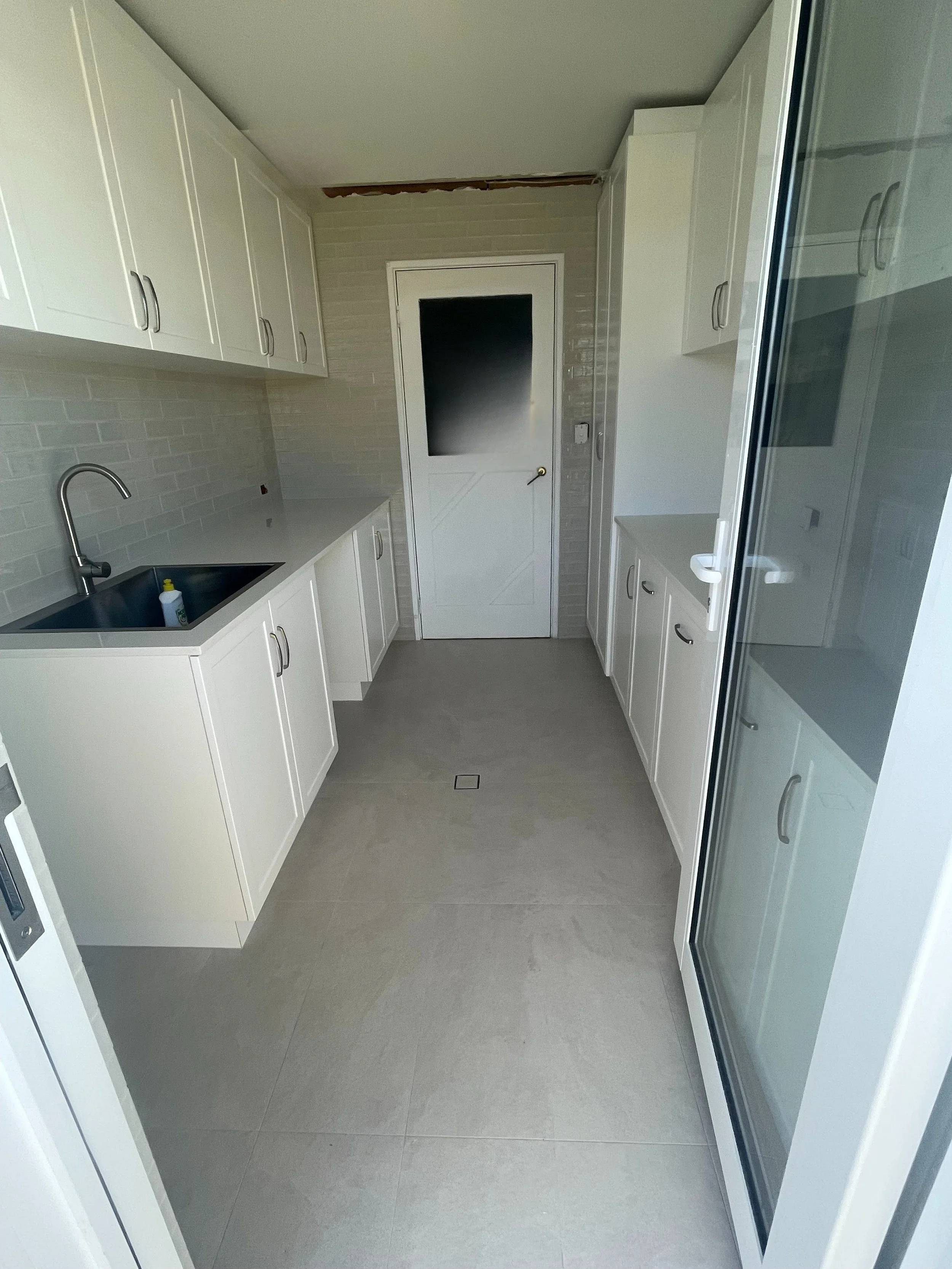 A narrow laundry room with white cabinets, a black sink with a faucet, a frosted glass door, and gray tiled floor.