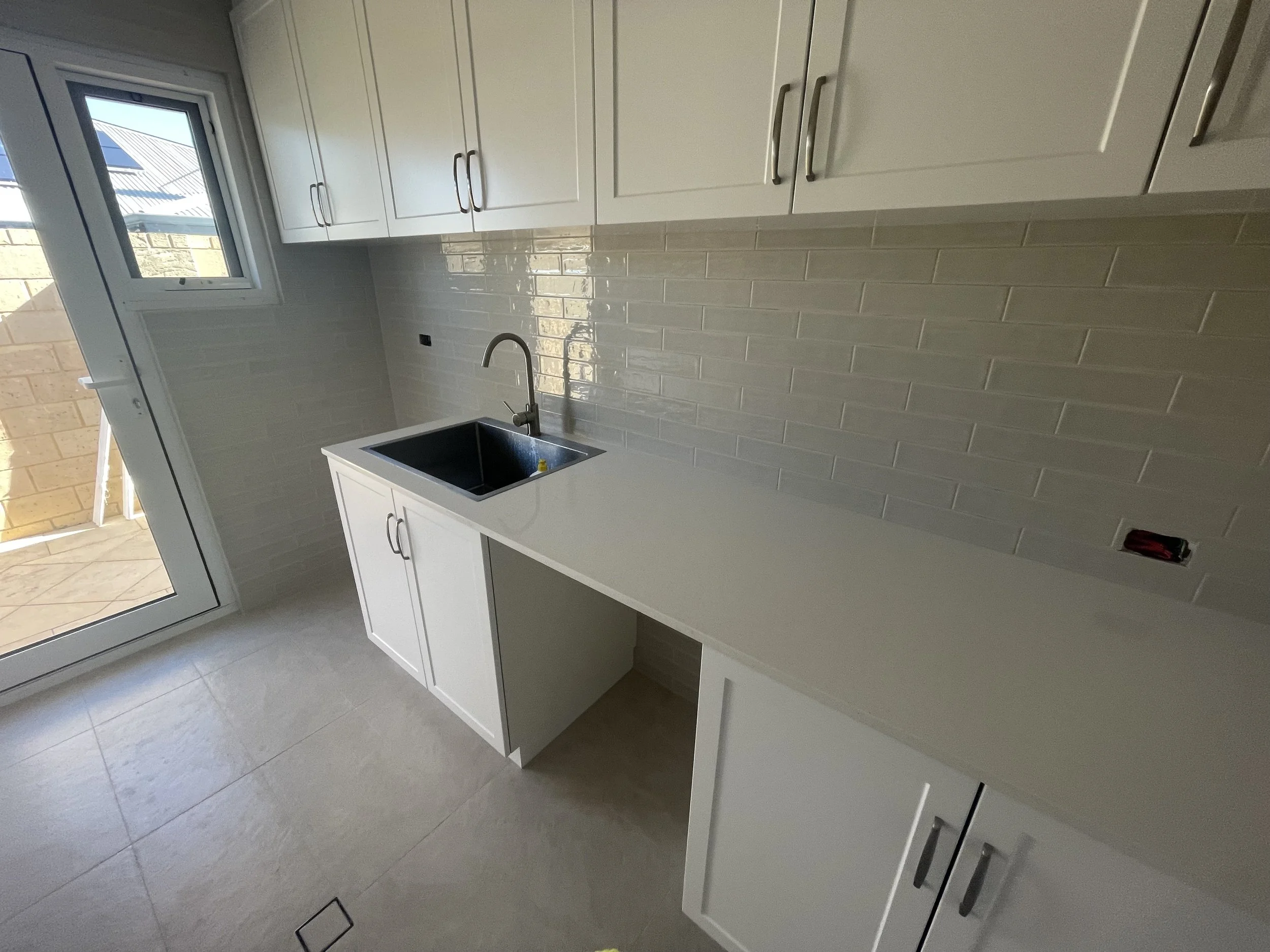 A kitchen scene with white cabinets, beige tiled backsplash, a black sink, and a faucet, with a door leading to an outdoor patio.