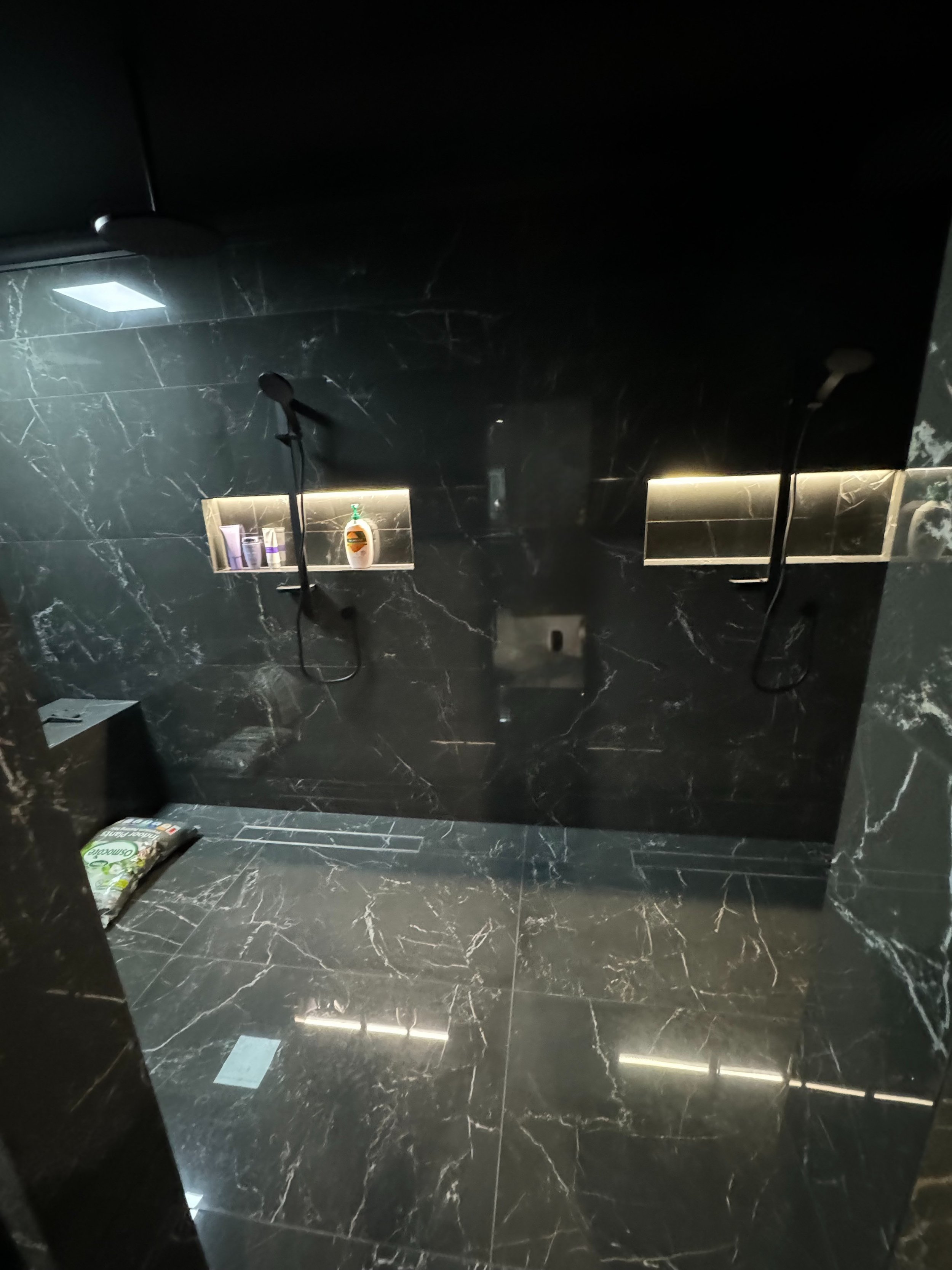 A sleek, modern shower room with black marble walls and floors, featuring two shower heads and built-in shelves with toiletries.
