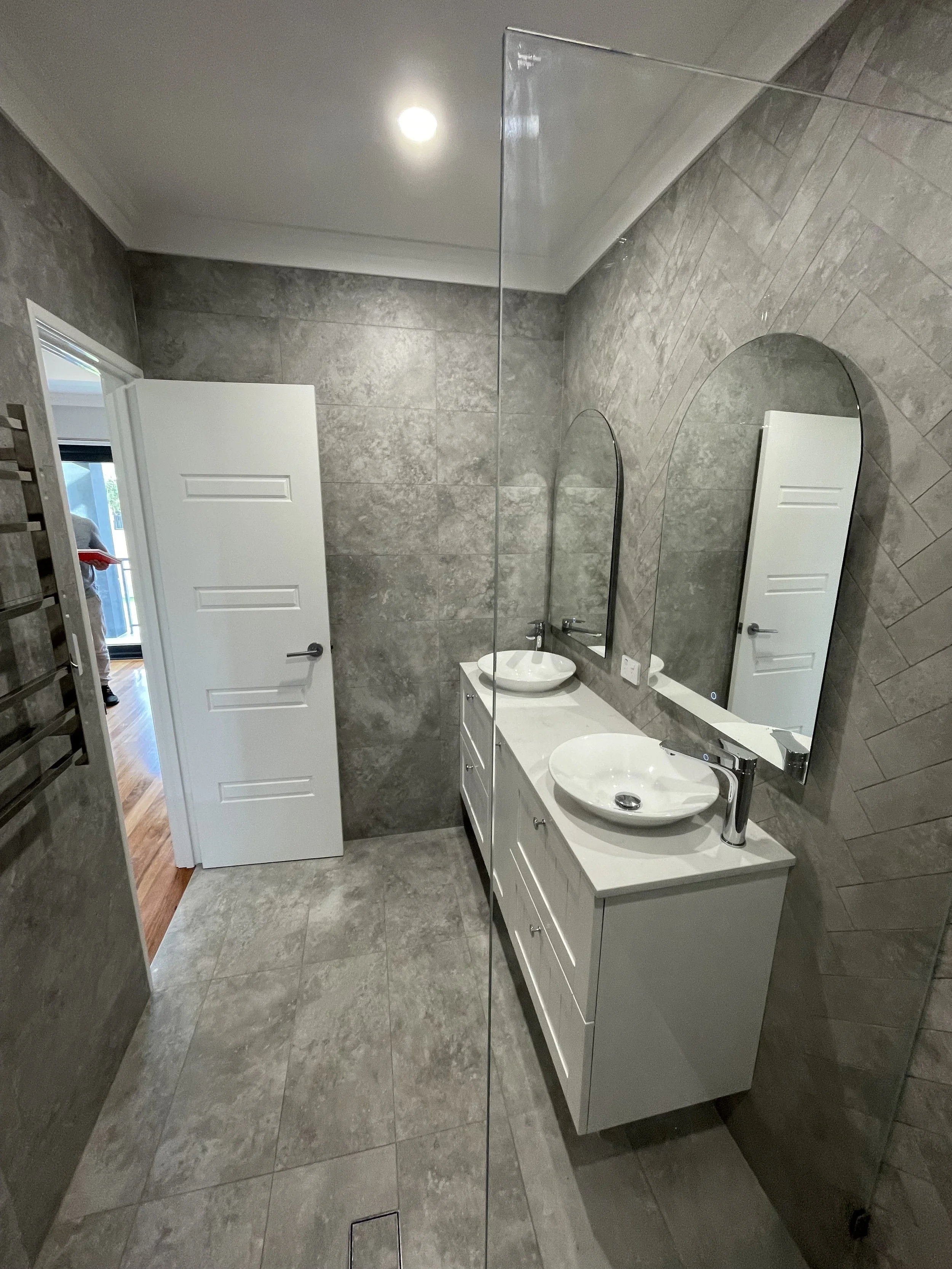 Modern bathroom with gray stone walls and floor, white vanity with two vessel sinks, large mirror, open door showing a room with hardwood flooring.