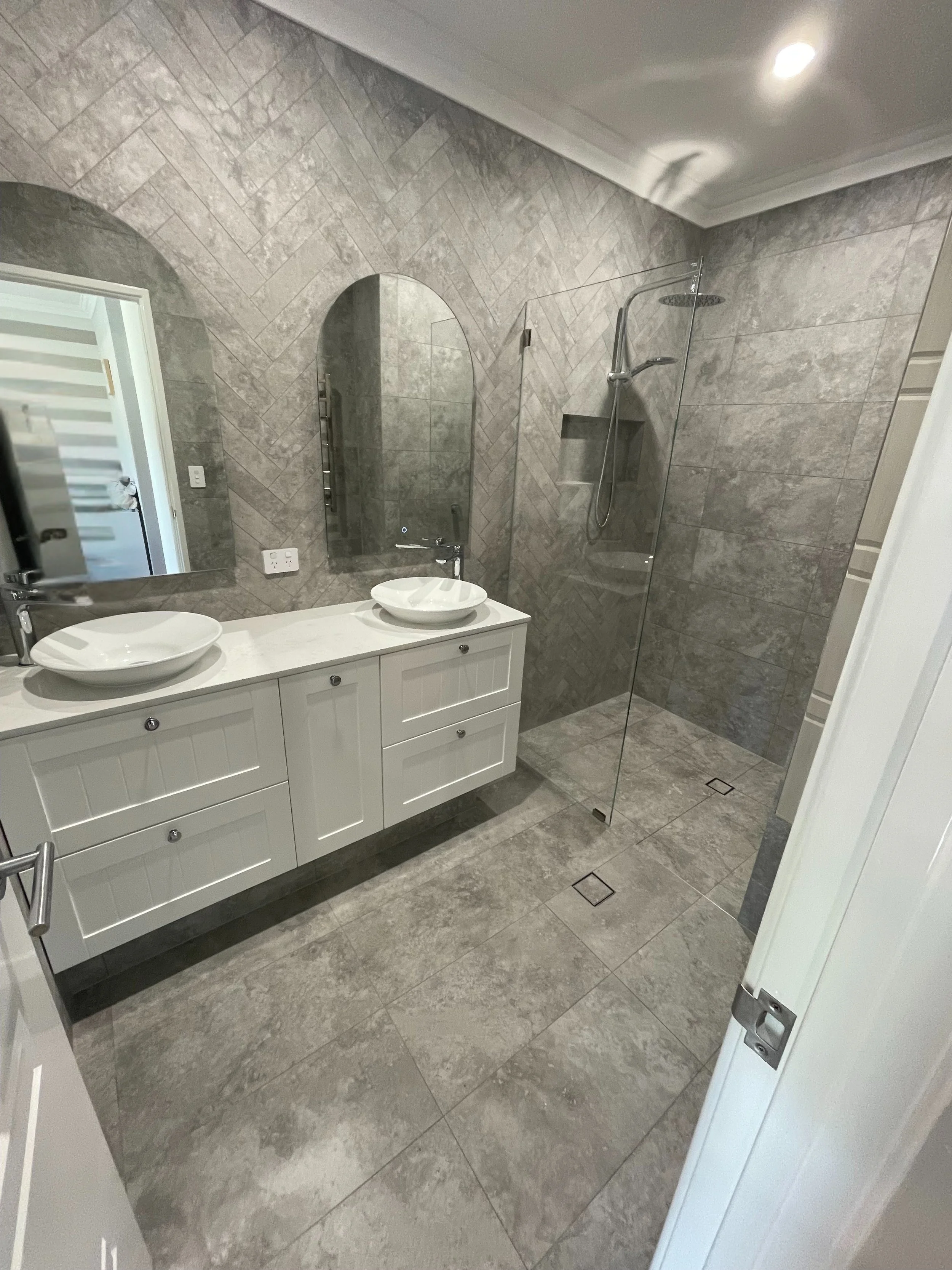 Modern bathroom with gray tiled walls and floor, double vanity with two sinks, oval mirrors, and a walk-in shower with a glass partition and built-in shelf.