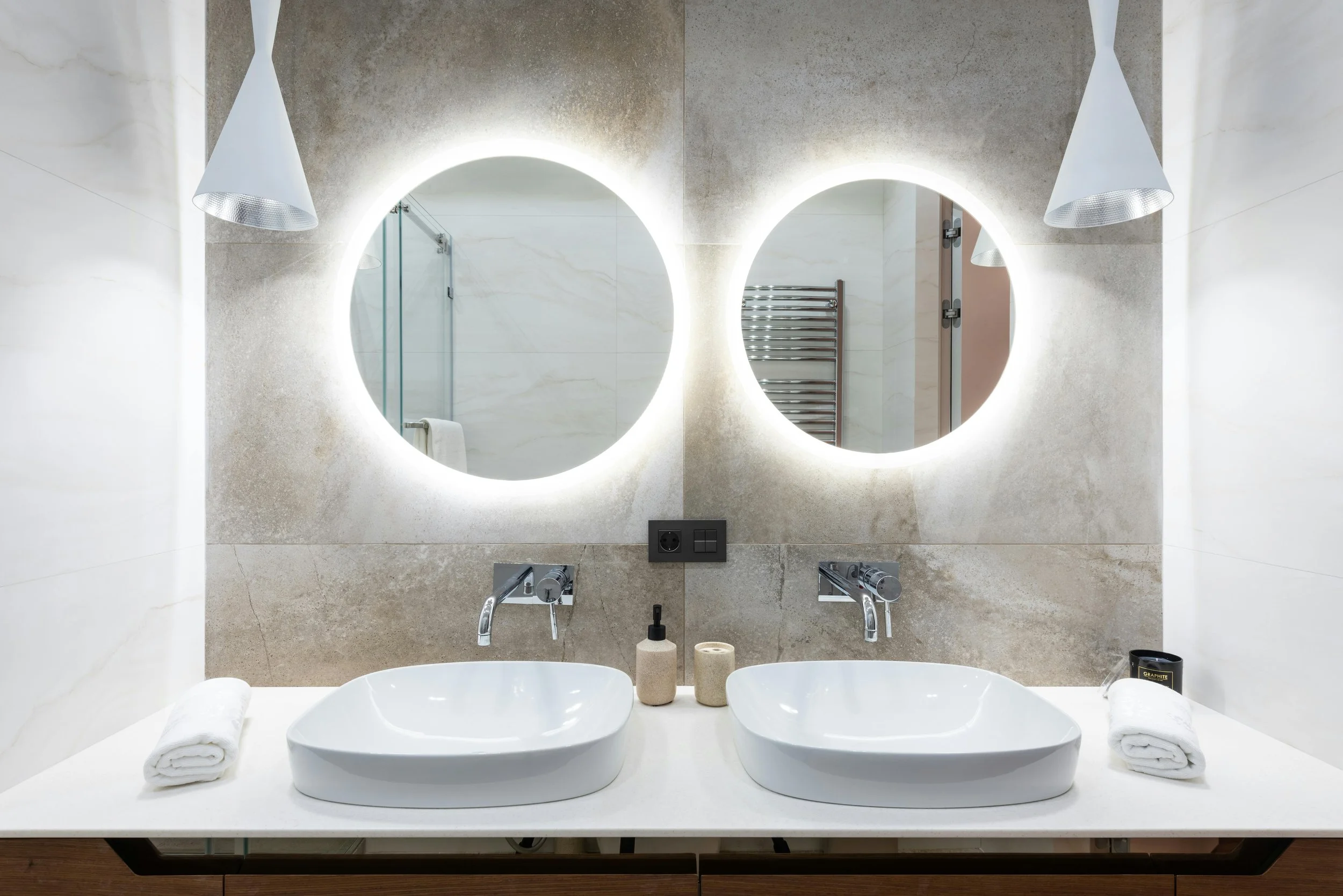 Modern bathroom with dual sinks, illuminated round mirrors, towels, soap dispenser, and wall-mounted faucets.