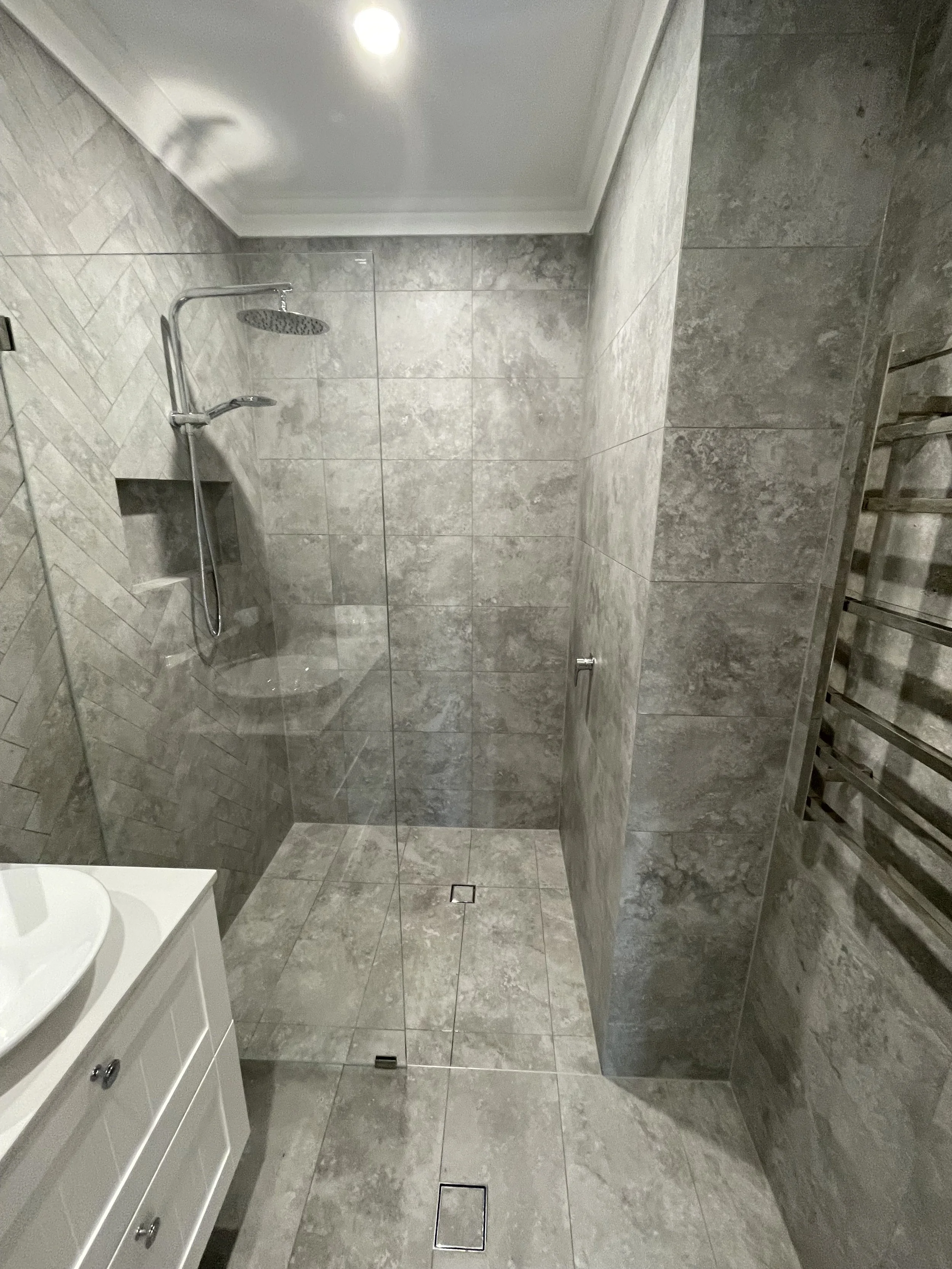 Gray tiled shower with a rain shower head, a small built-in shelf, a glass partition, a towel radiator on the right wall, and a white vanity with a vessel sink in the corner.