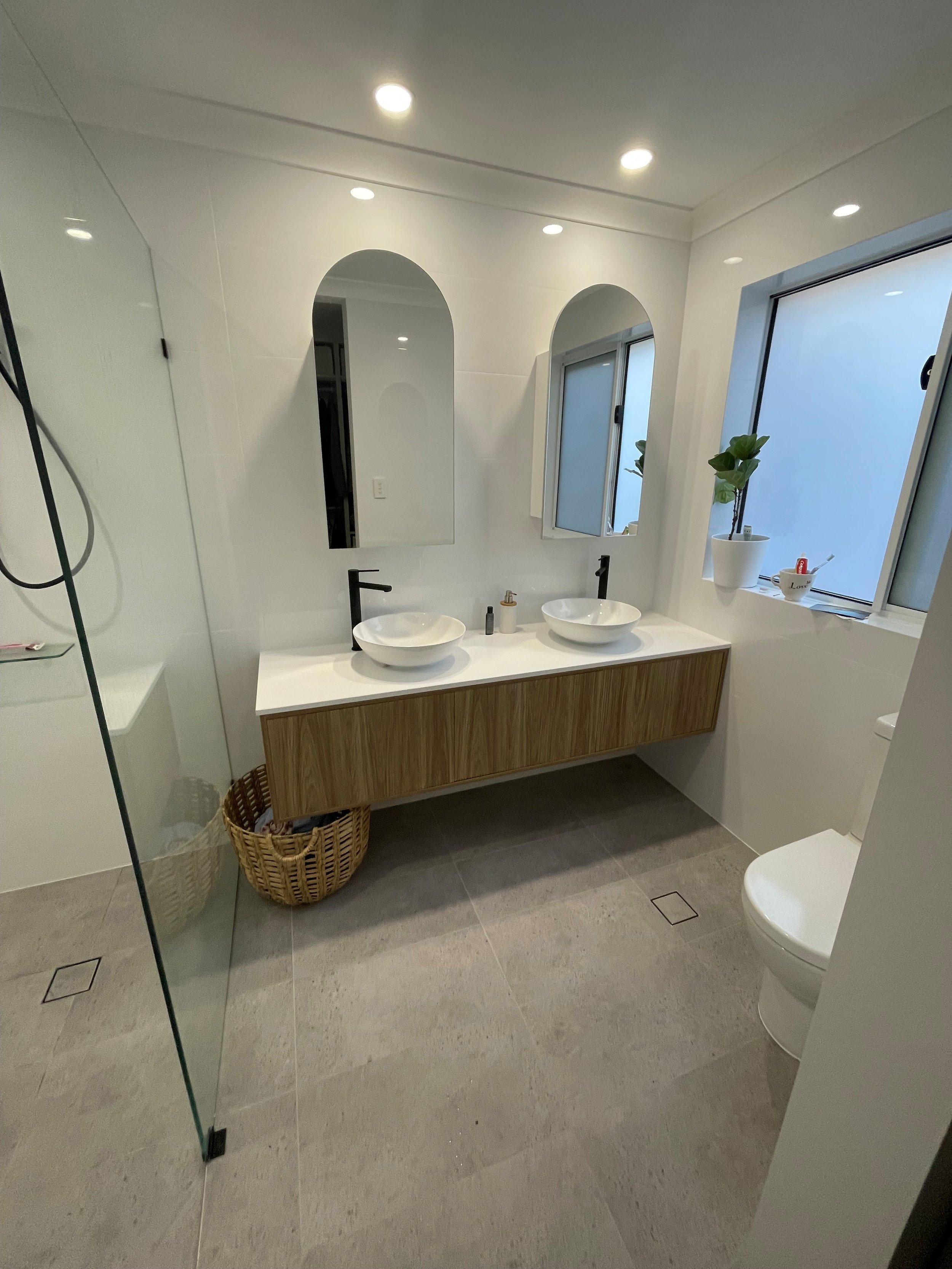 Modern bathroom with double vanity, two vessel sinks, black faucets, two arched mirrors, frosted window, potted plant, wicker basket, and glass shower enclosure.
