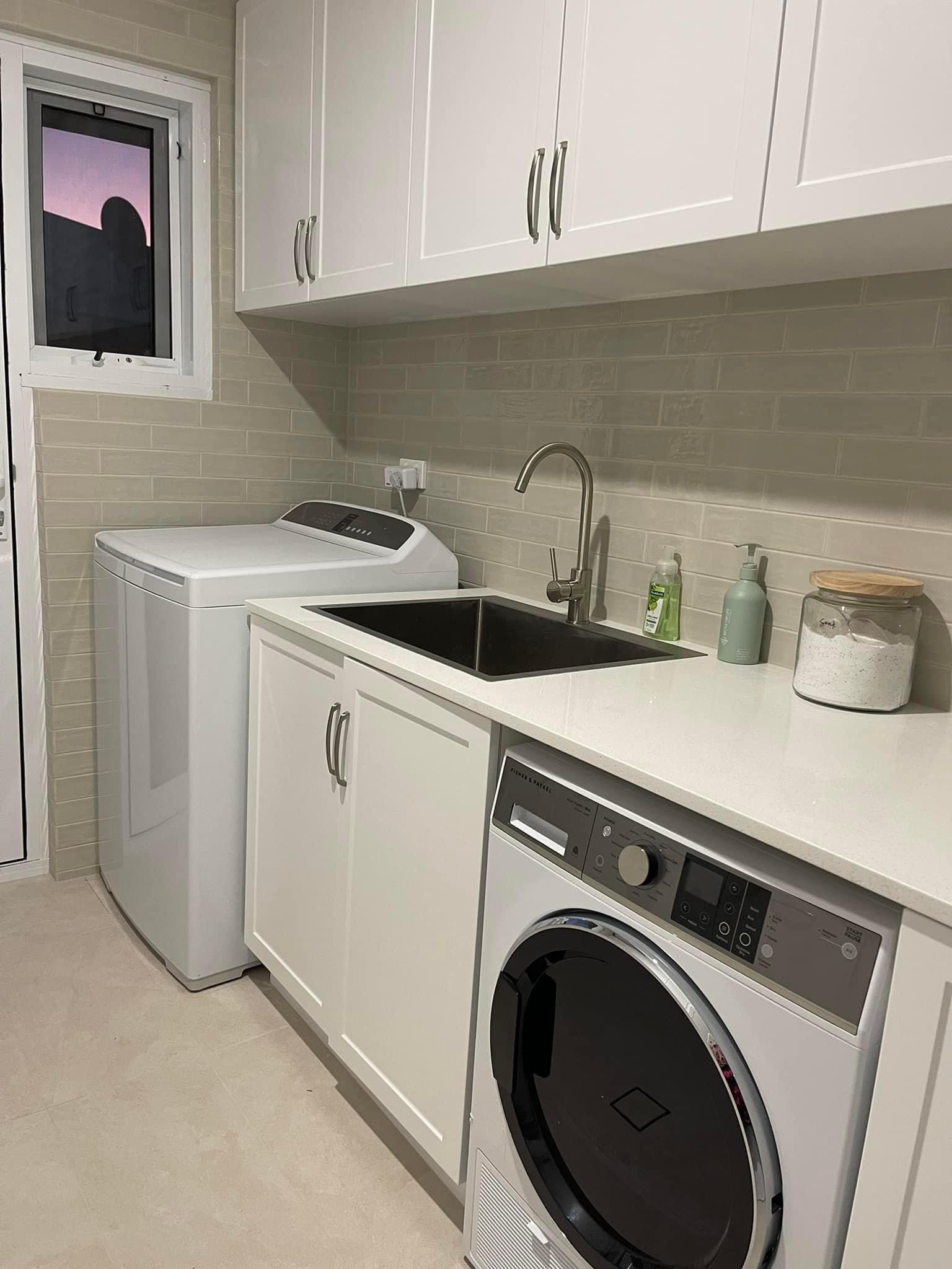 A laundry room with a washing machine, a sink, white cabinets, and countertop, with soap and sanitizer bottles, a jar of detergent, and a window showing a sunset or sunrise.