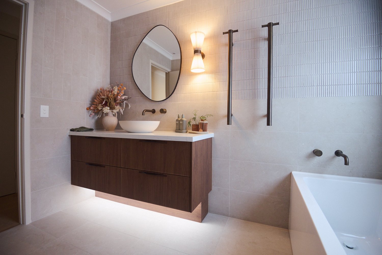 Modern bathroom with a floating dark wood vanity, white oval sink, round mirror, wall-mounted light fixture, decorative flowers, and a bathtub with wall-mounted faucet.