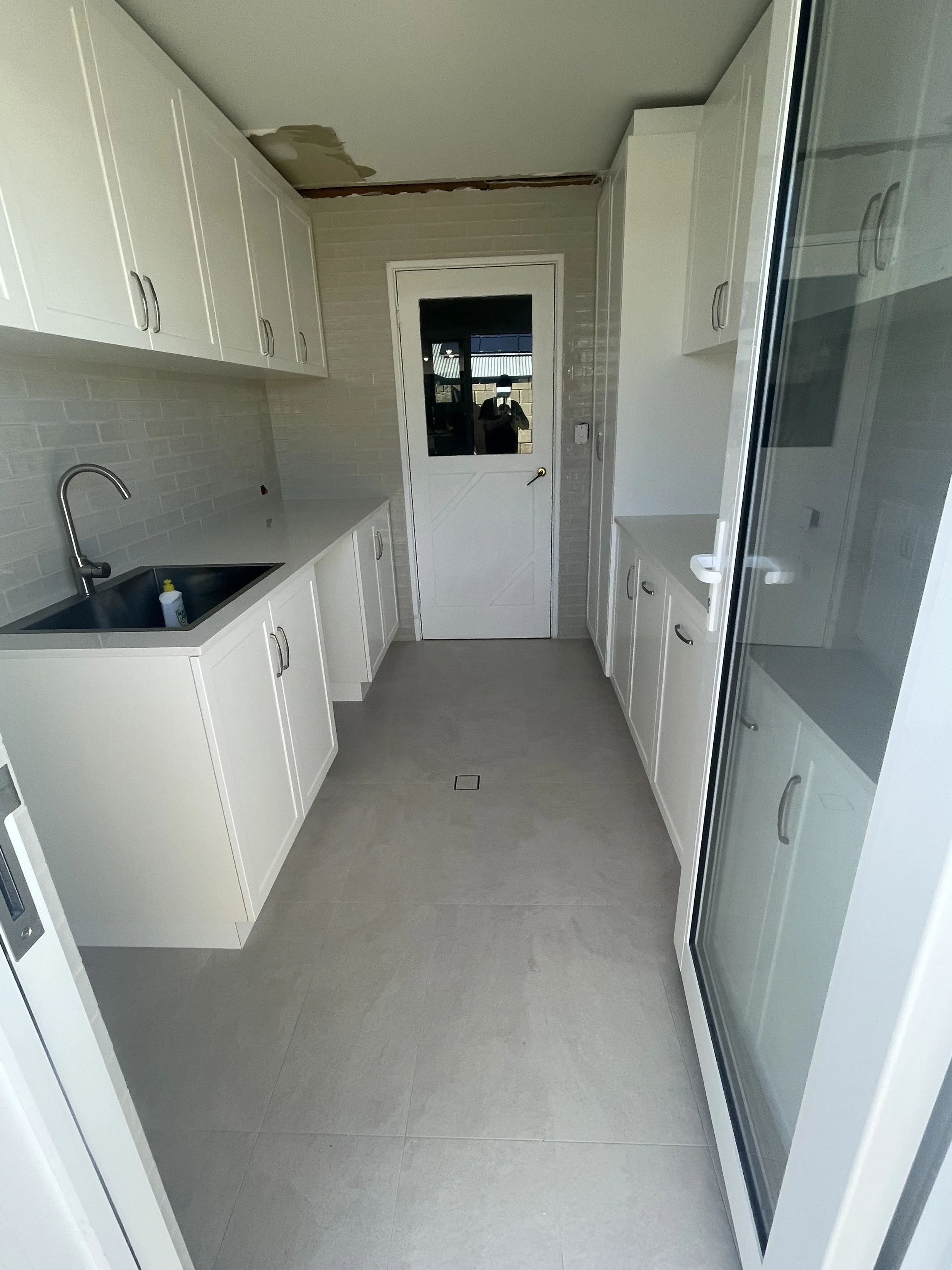 Clean, modern laundry or utility room with white cabinetry, gray tiled flooring, a small sink, and a door with glass panel leading outside. Some ceiling repair area visible.