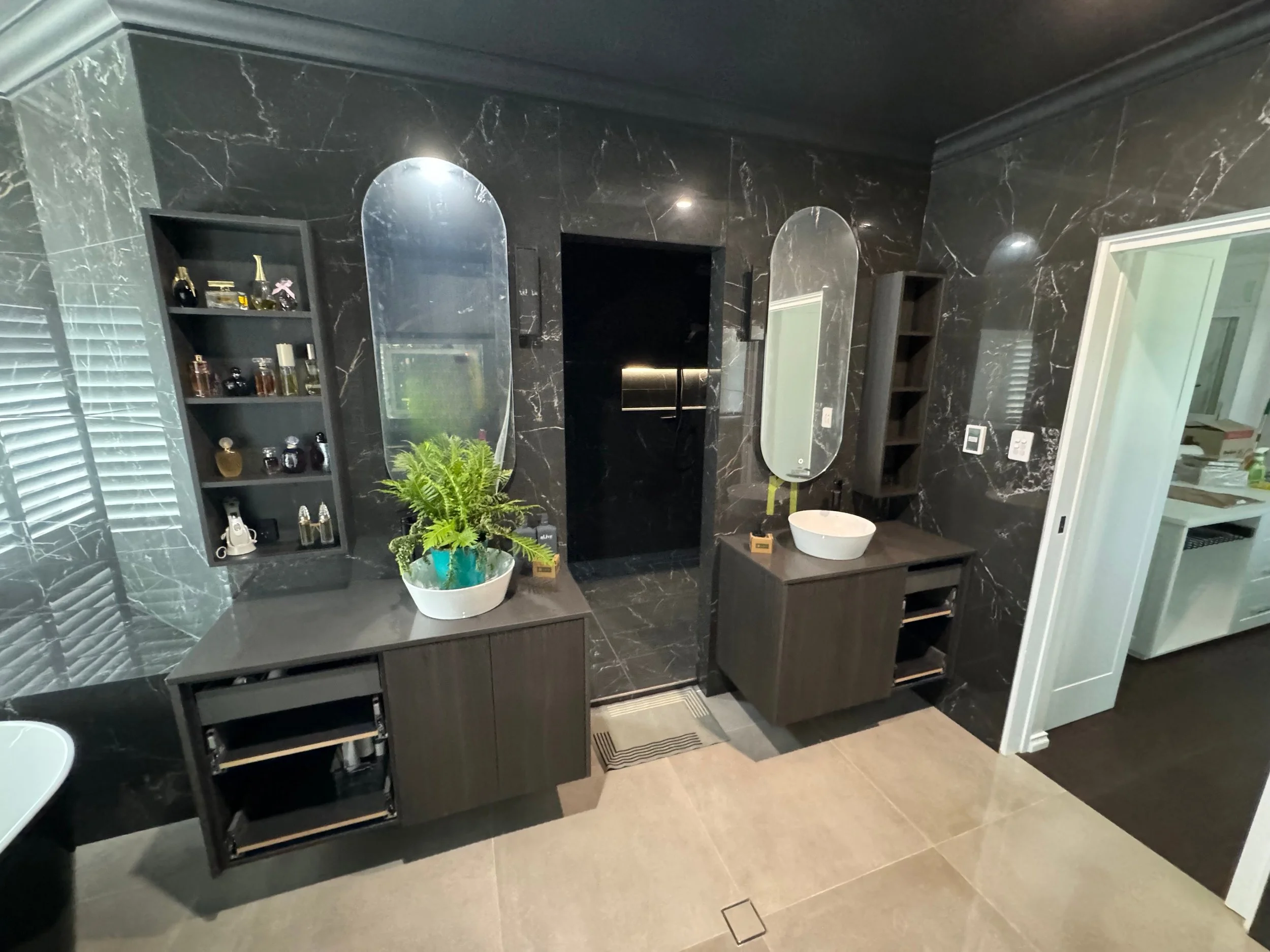 Bathroom with black marble walls, two dark wood vanities with oval mirrors, one with a white vessel sink, a potted fern on the left vanity, small black shelving units, and a laundry area visible through a sliding door on the right.