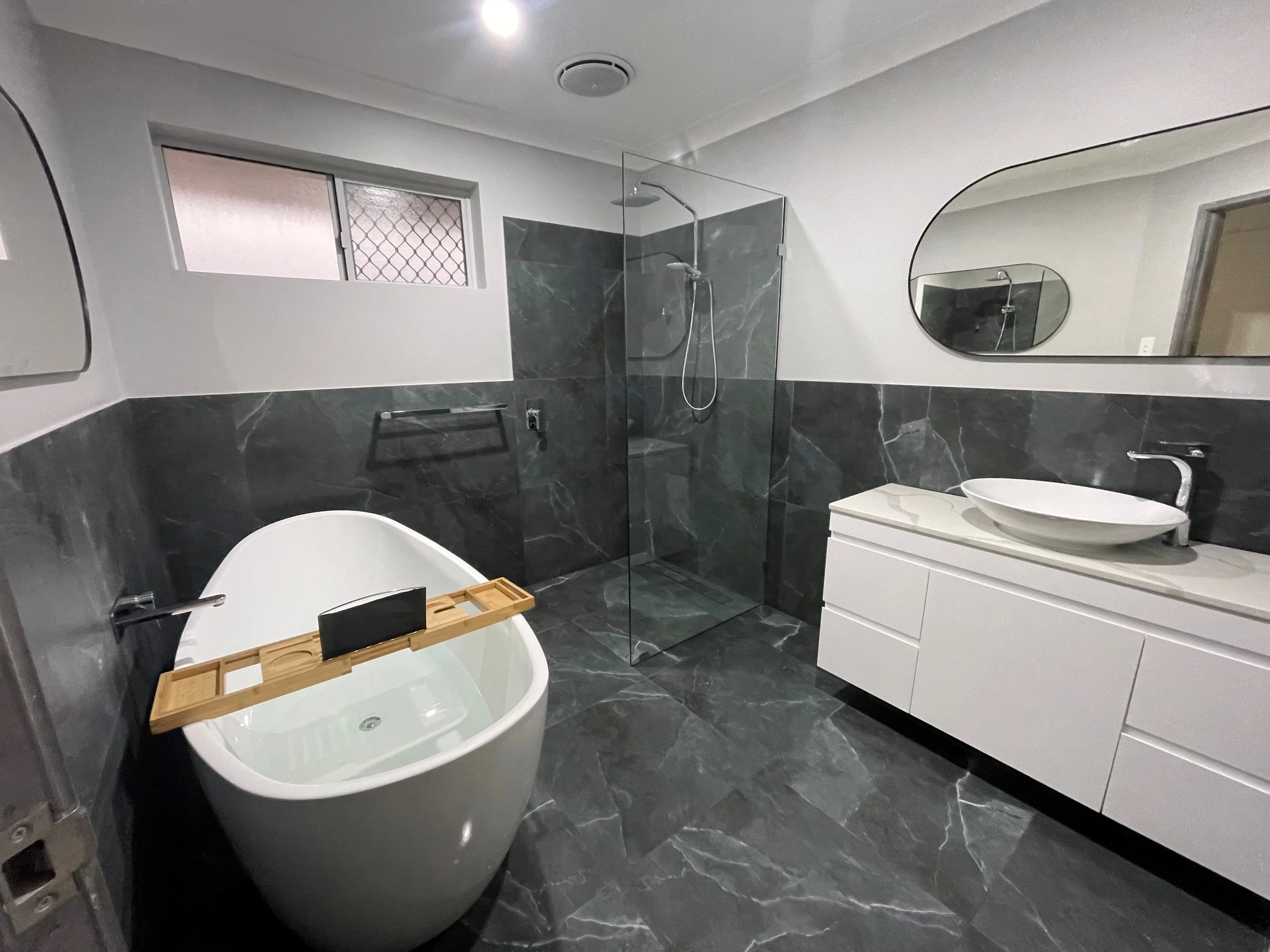 Modern bathroom with dark gray marble floors and half-wall tiles, white bathtub, glass shower enclosure, white vanity with oval vessel sink, oval mirror, and frosted and patterned windows.